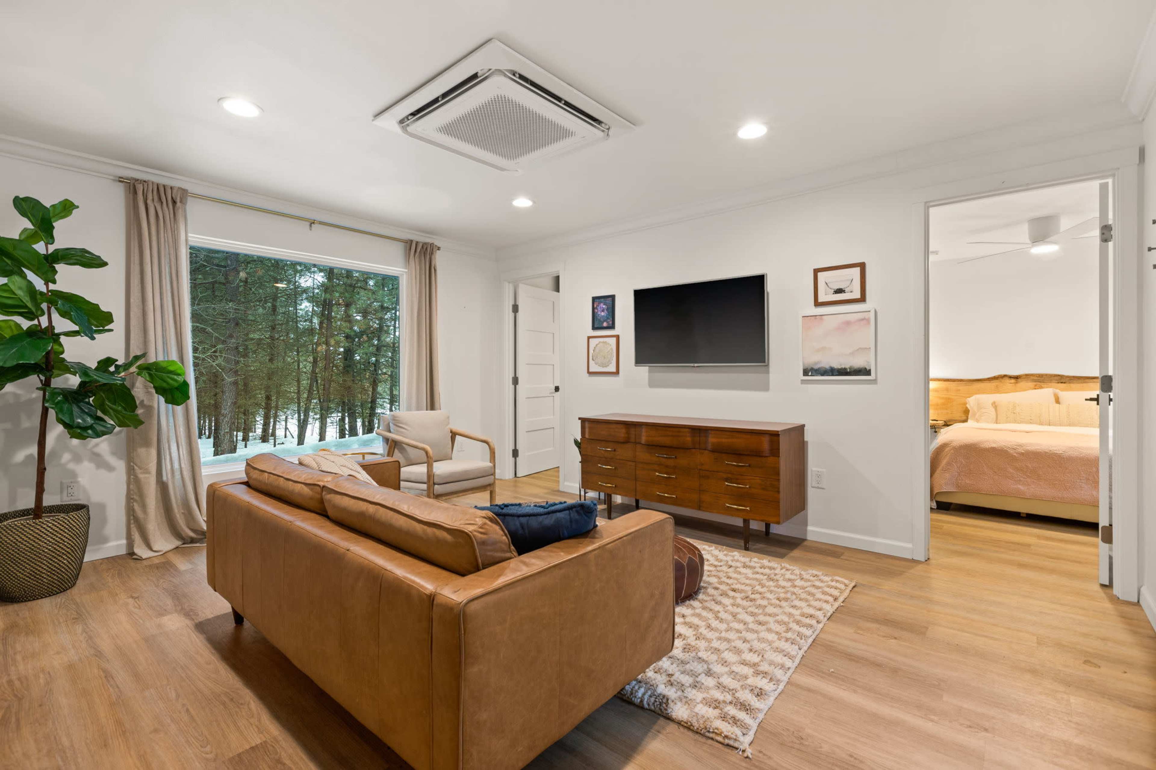 The living room features a leather sofa, a TV mounted on the wall, and a large window overlooking a forested area.