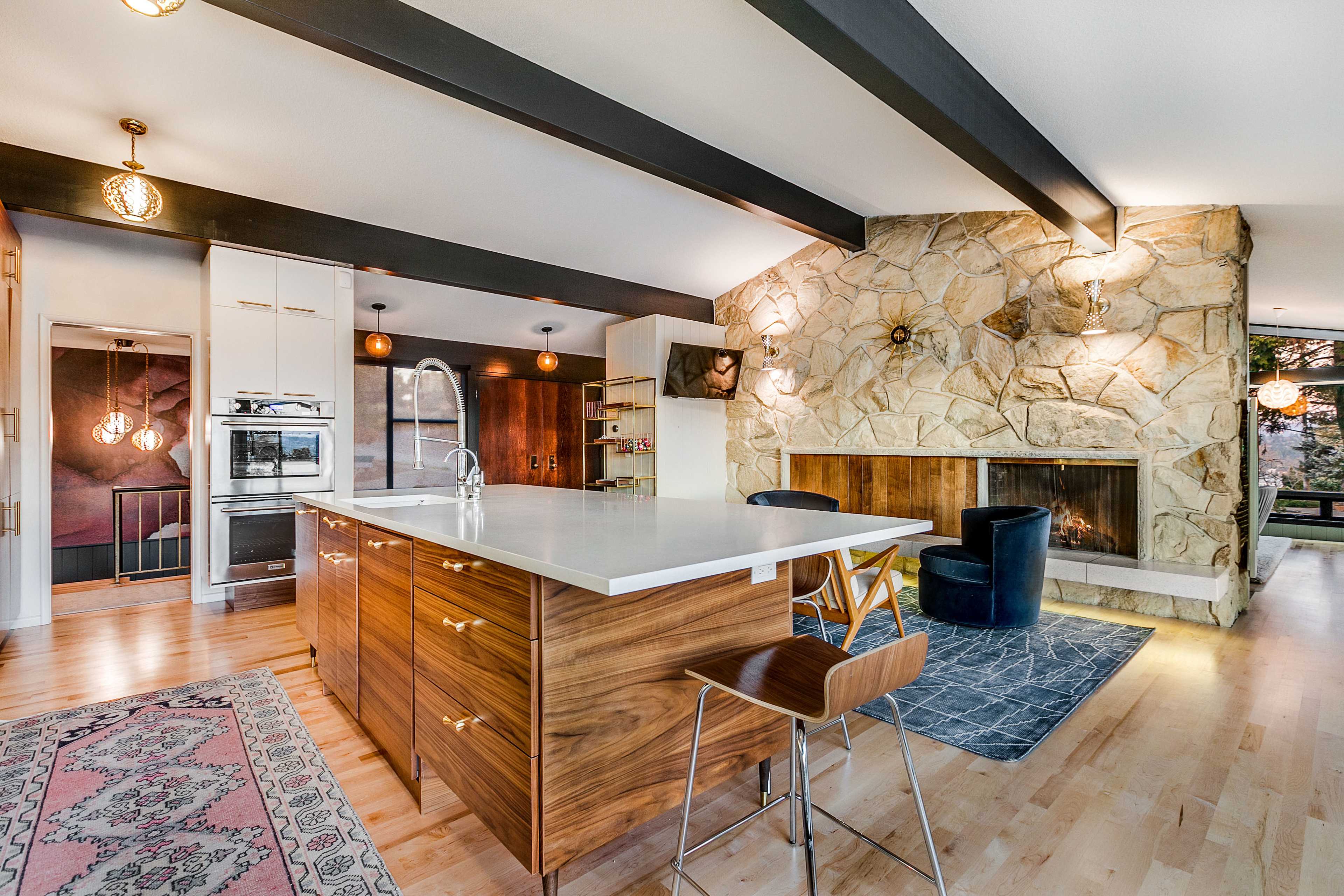 A modern kitchen features a central island with wooden cabinetry, a stone fireplace, and exposed ceiling beams.
