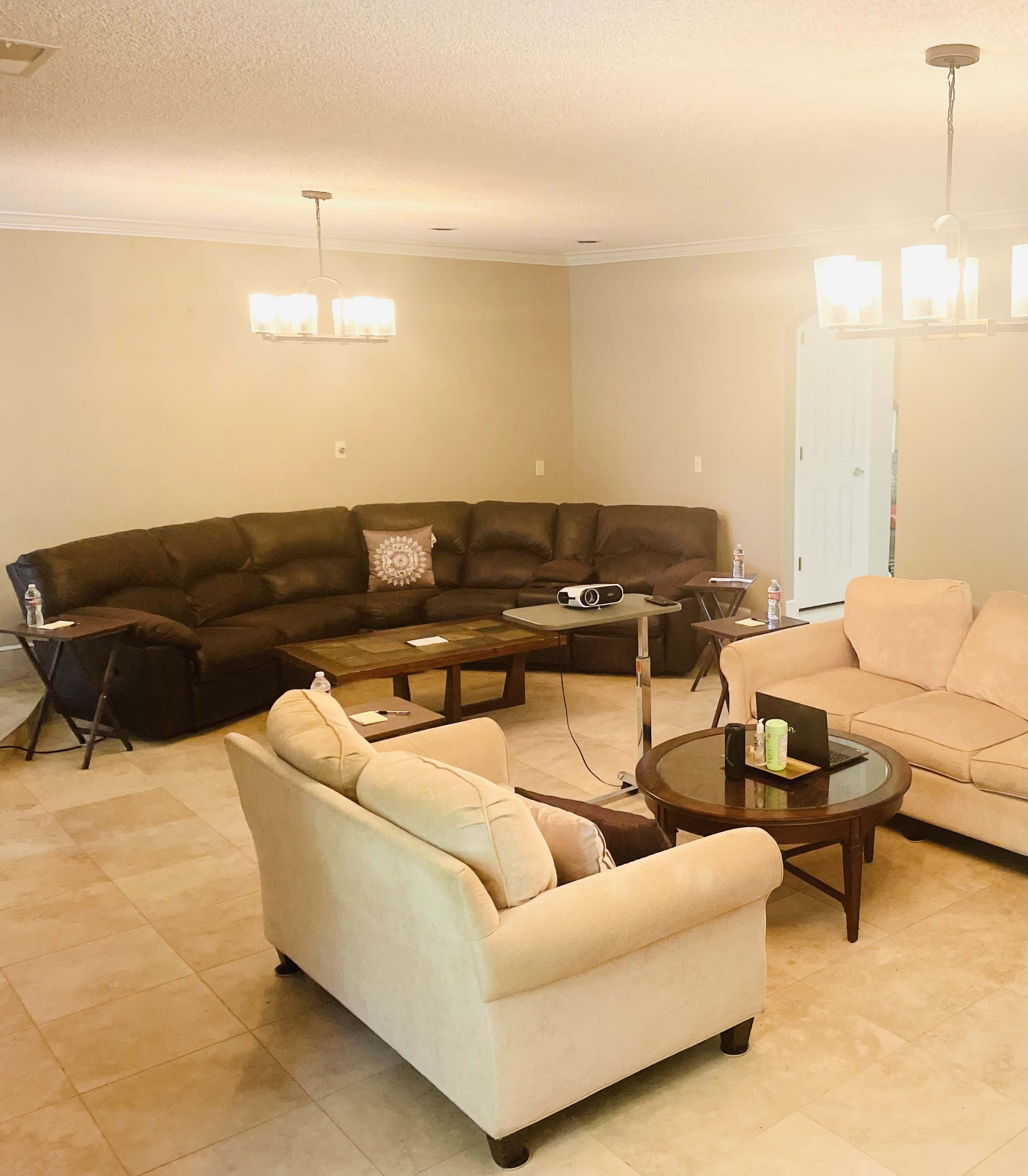 The image shows a spacious living room with a large brown sectional sofa, two cream-colored armchairs, a wooden coffee table, and a projector set up on a stand.