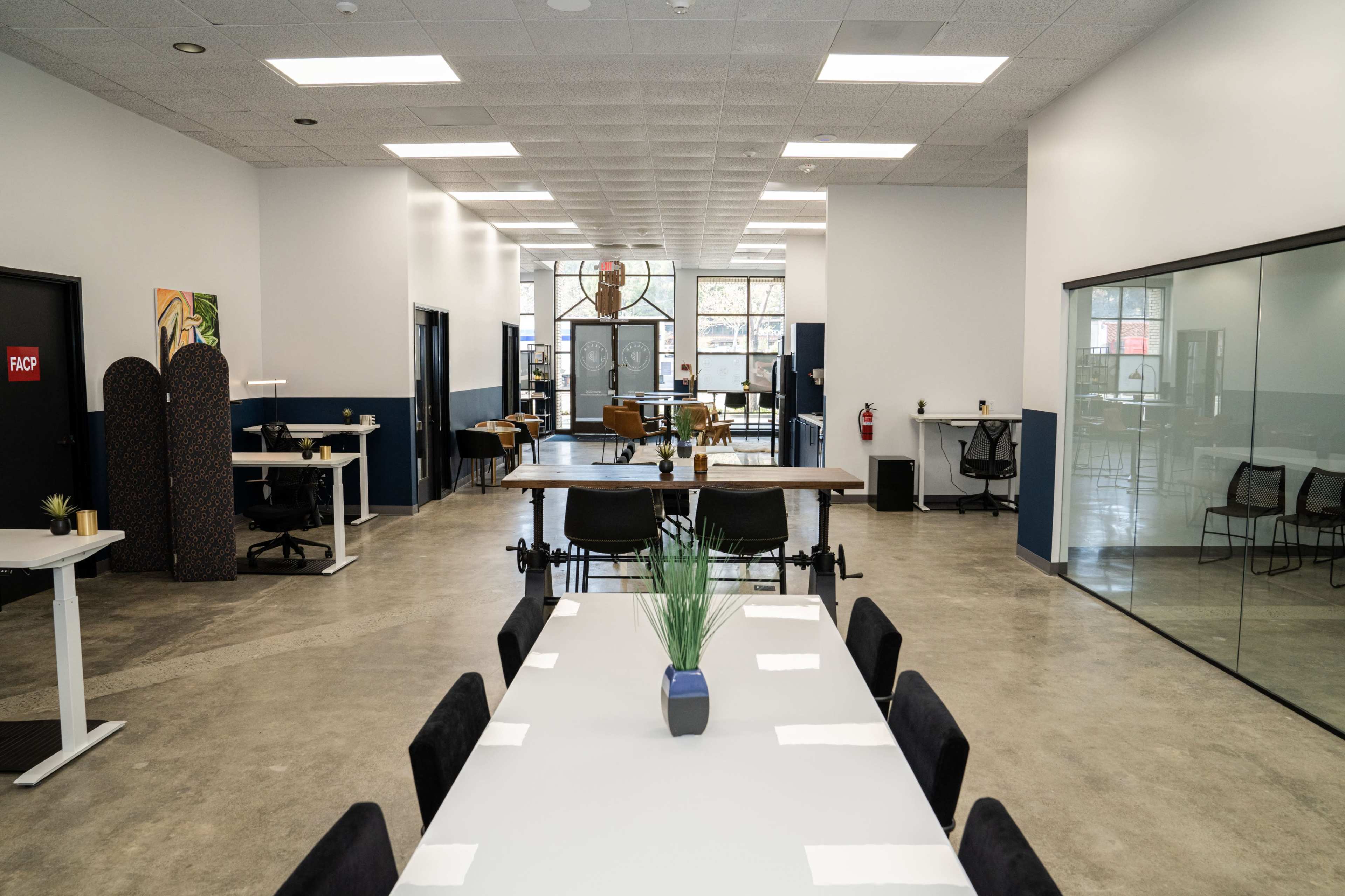 A modern office space with a large communal table, individual desks, and a glass-walled meeting room.