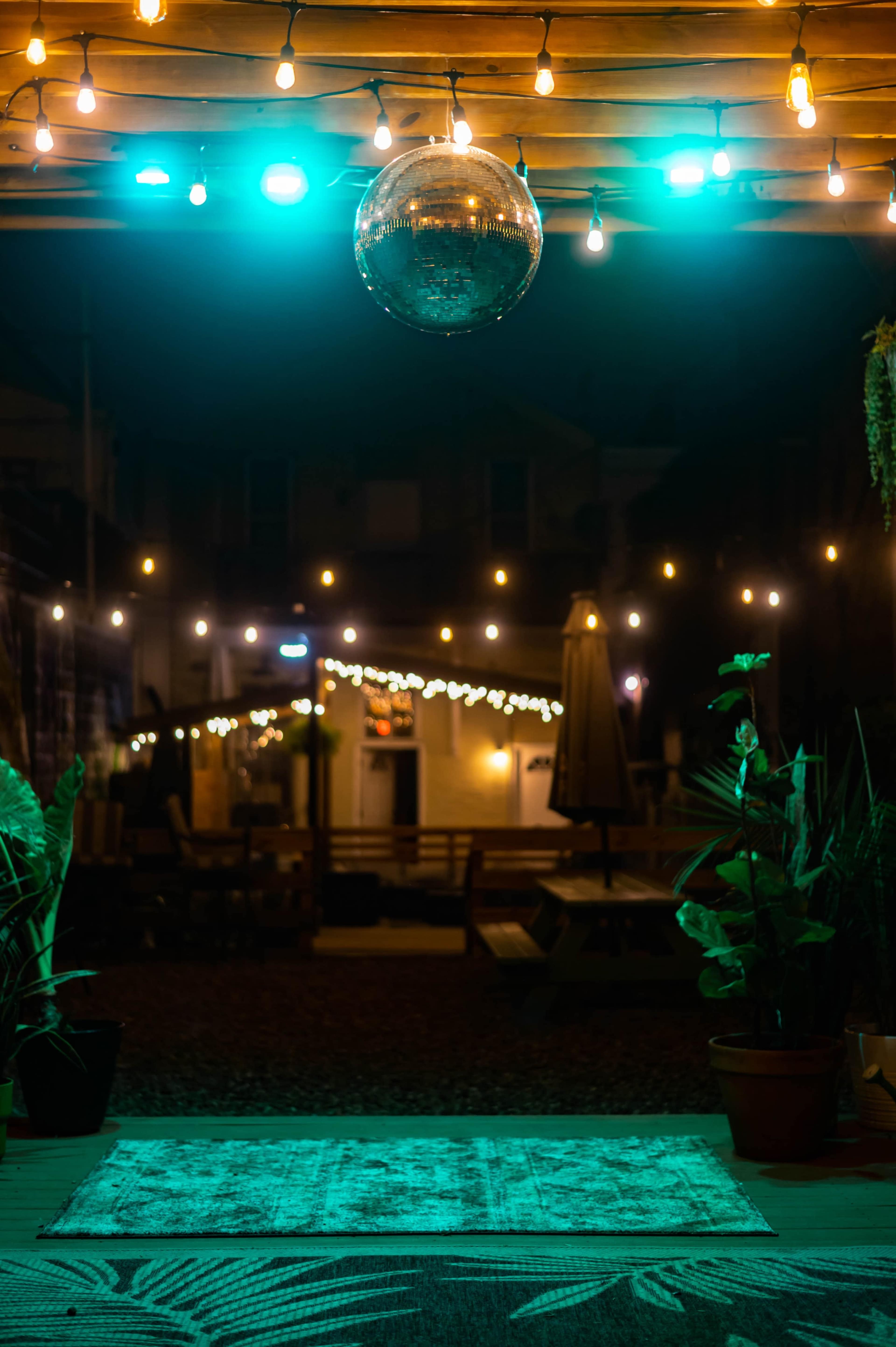 A dimly lit outdoor area featuring a disco ball hanging from a ceiling, with string lights illuminating the space and potted plants in the foreground.