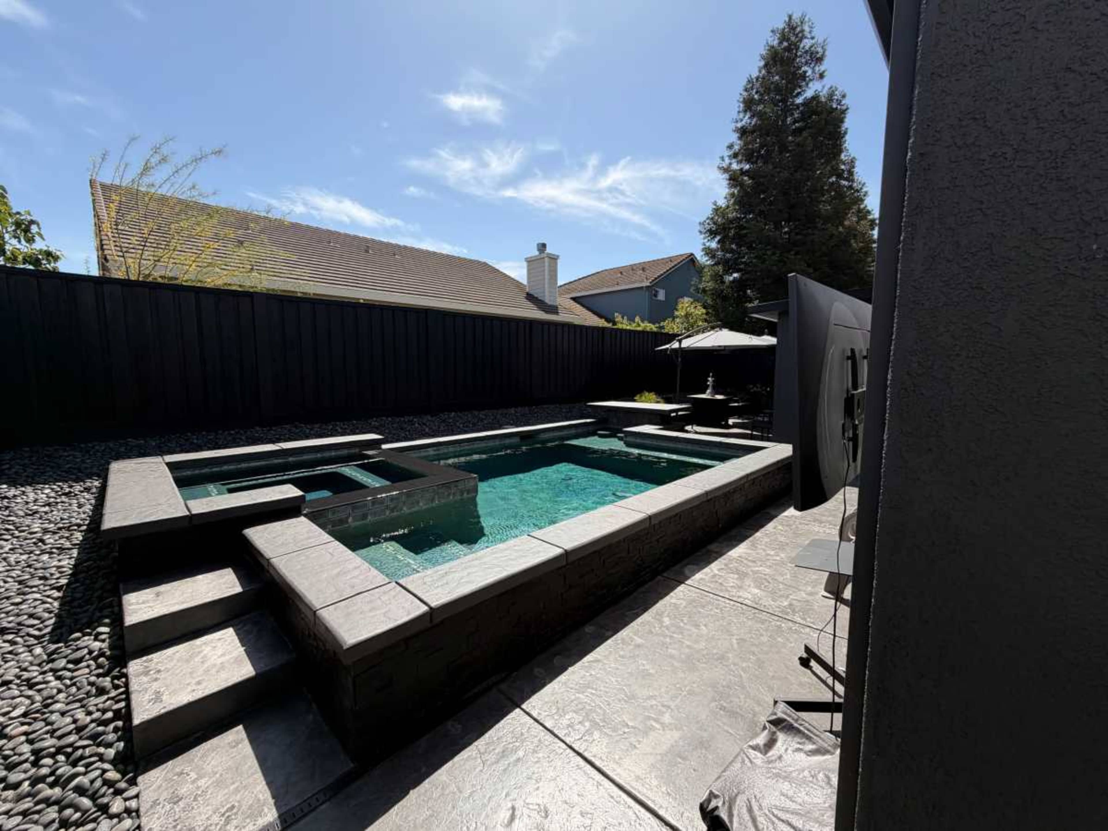 The image shows a modern backyard with a rectangular pool surrounded by gray stone steps and landscaping, bordered by a black fence and houses in the background.