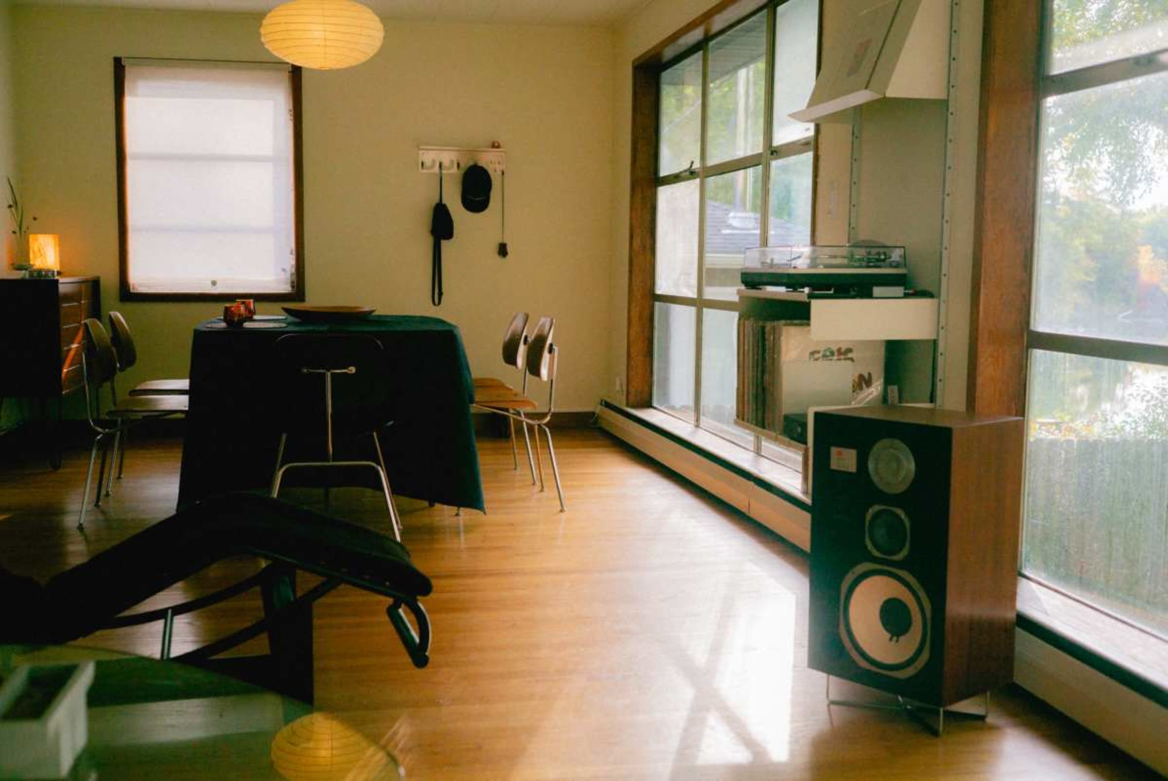 A spacious room features a dining table with chairs, a record player on a shelf, and a vintage speaker next to large windows.