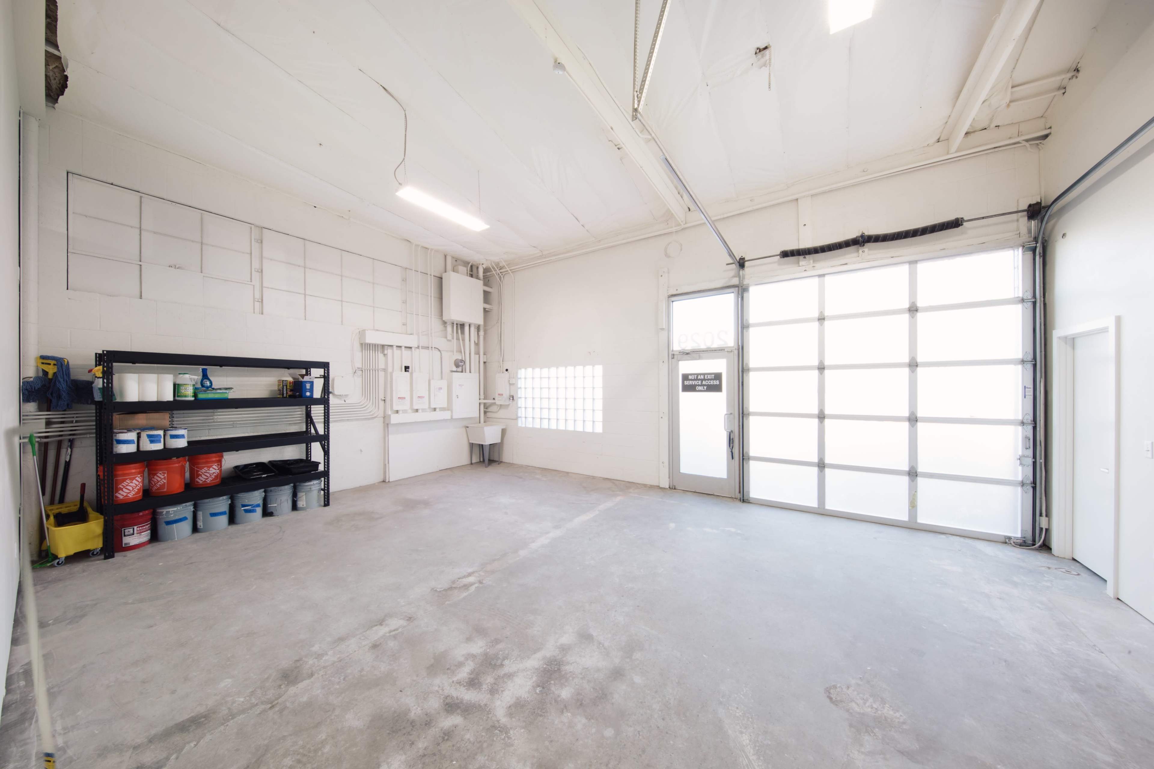 An empty garage with a concrete floor, a large roll-up door, and shelves filled with various storage containers.