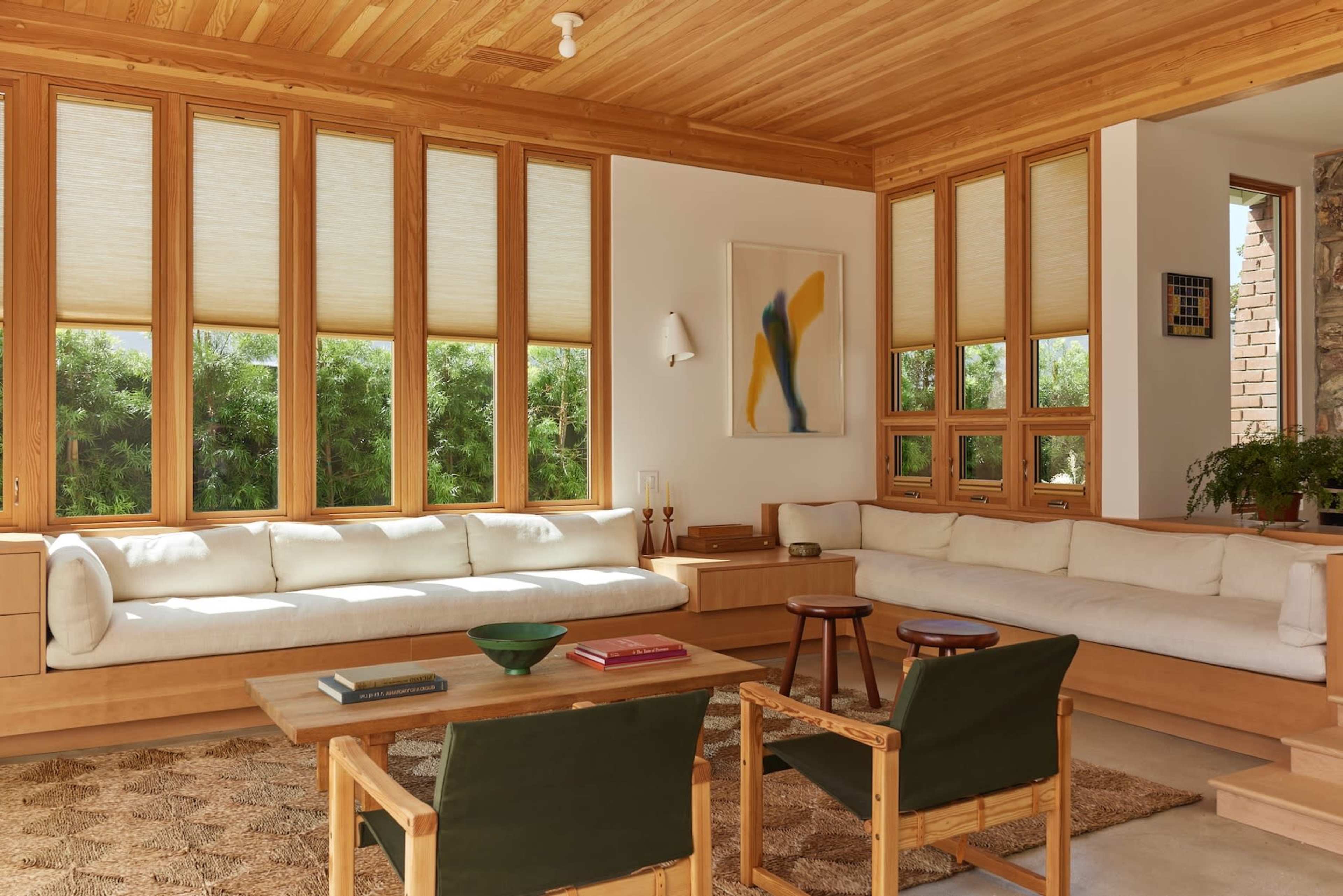 The living room features large windows, wooden walls and ceiling, and minimalist furniture arranged around a central table.