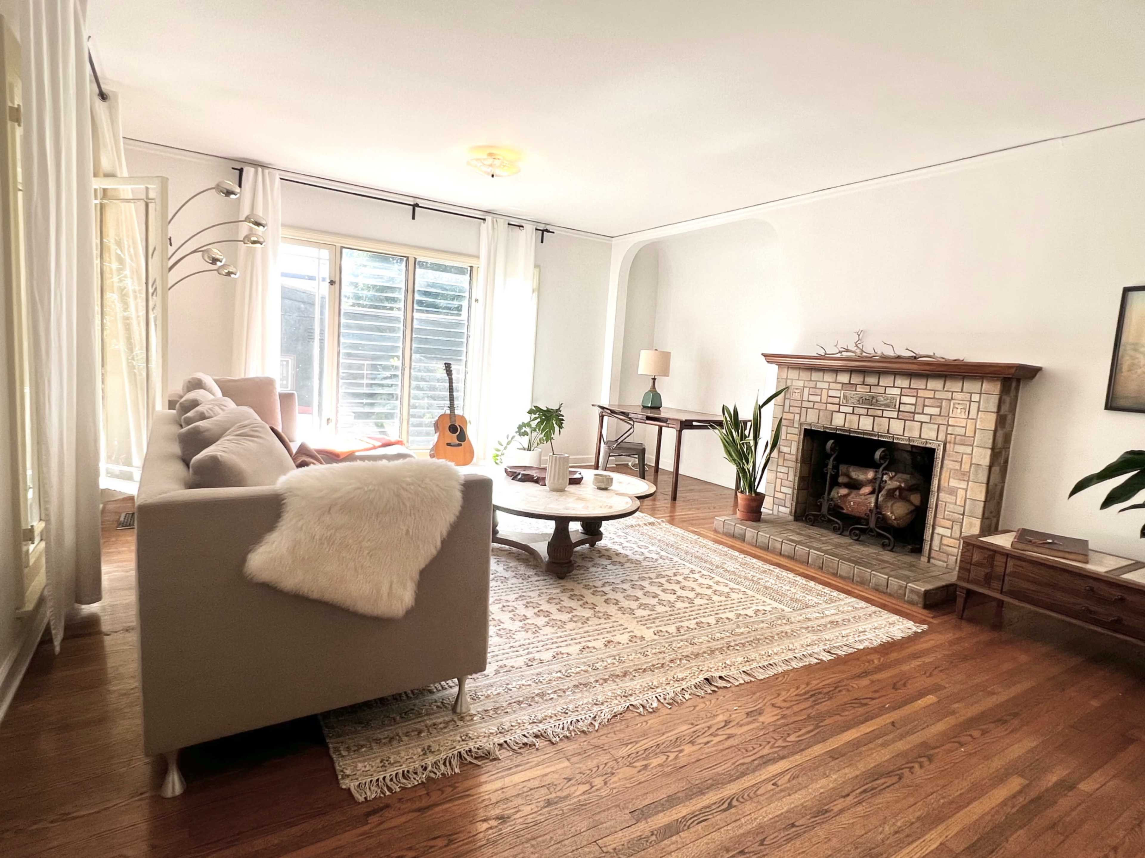 A cozy living room features a gray sofa, a wooden coffee table, a fireplace with a stone surround, and large windows allowing natural light.