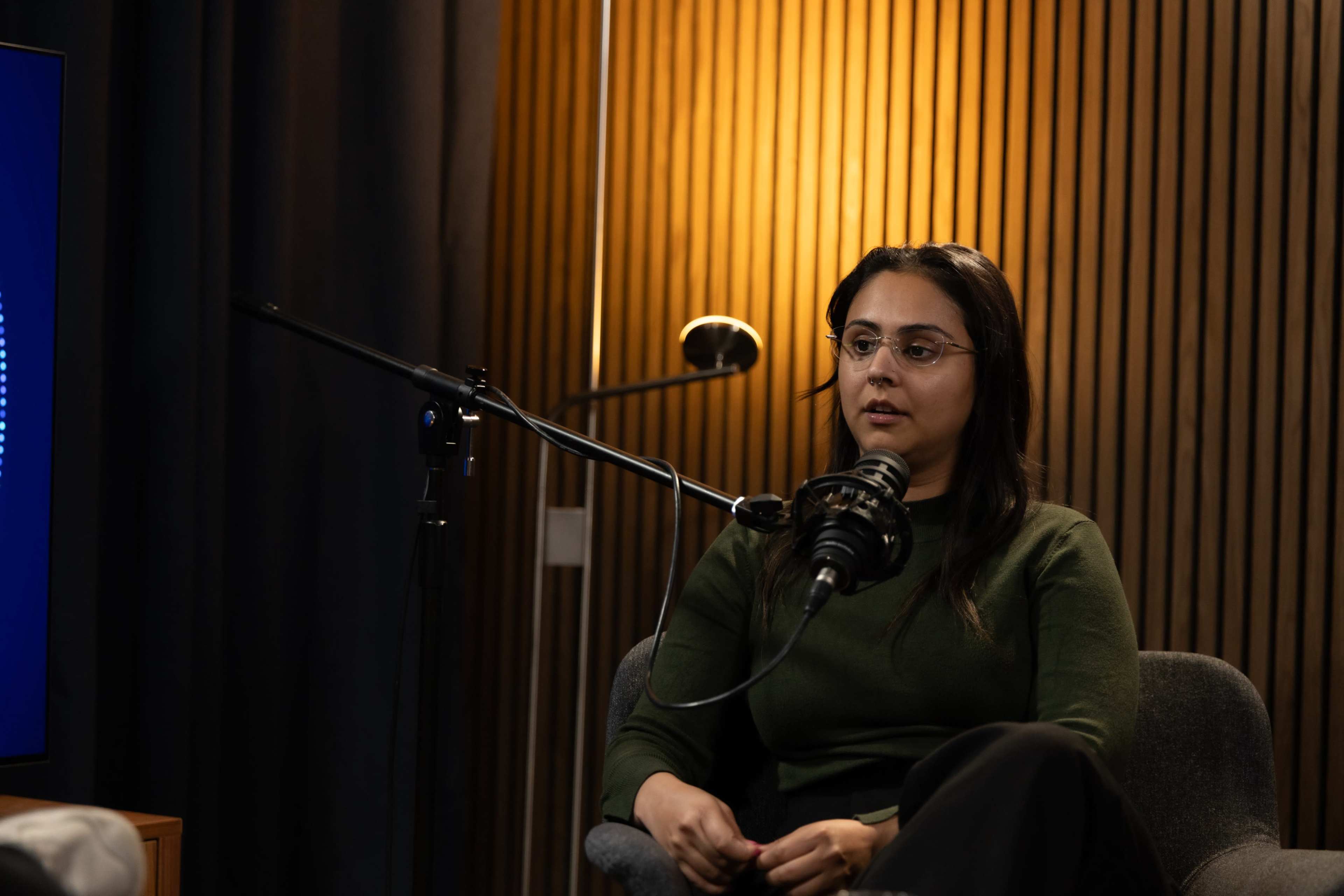 A woman sits in a chair with a microphone in front of her, surrounded by wooden paneling and soft lighting.