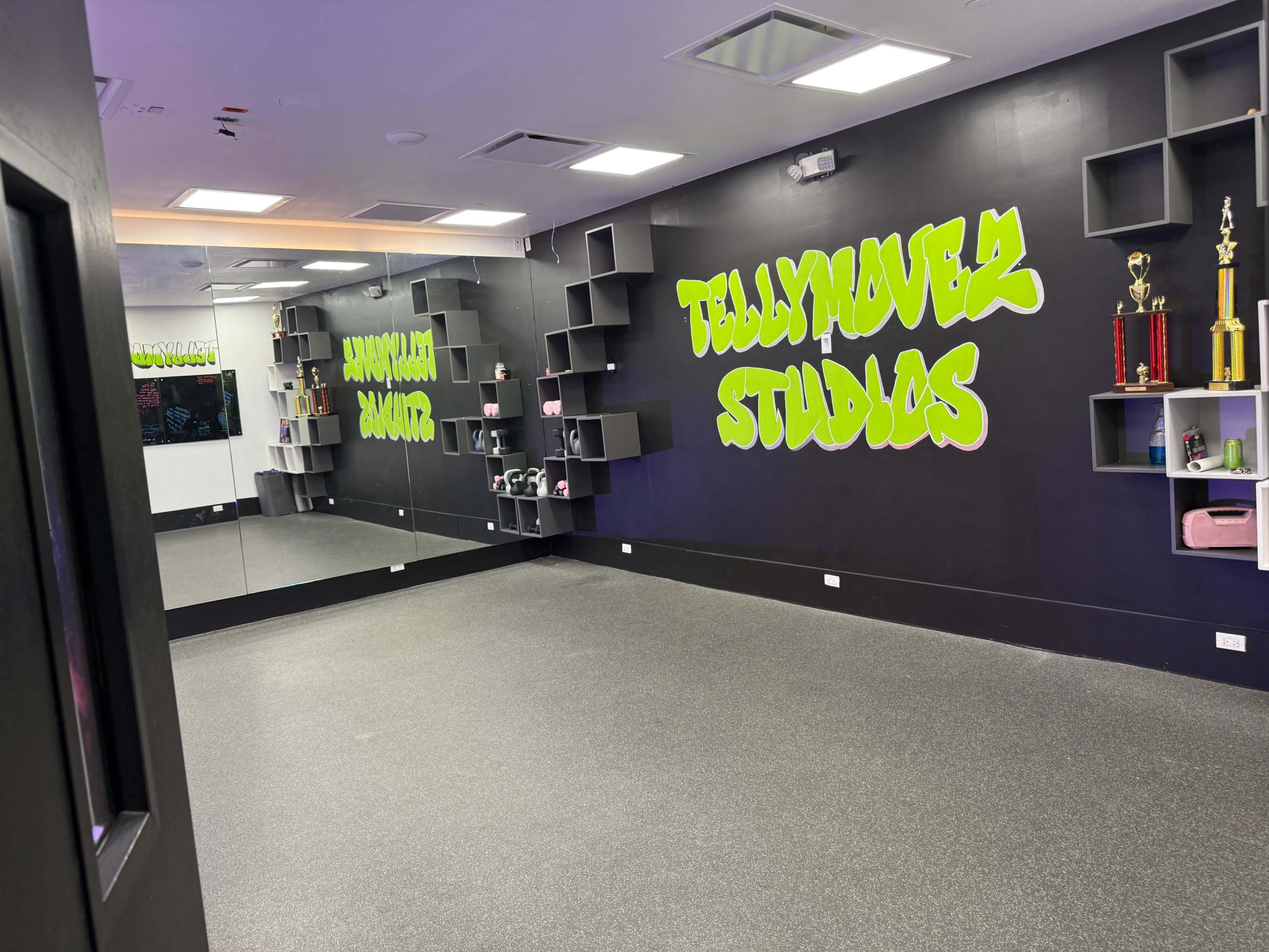 The image shows a room labeled "TellyMover Studios" with a black wall, green graffiti-style lettering, a mirrored wall, and various trophies and shelves on display.