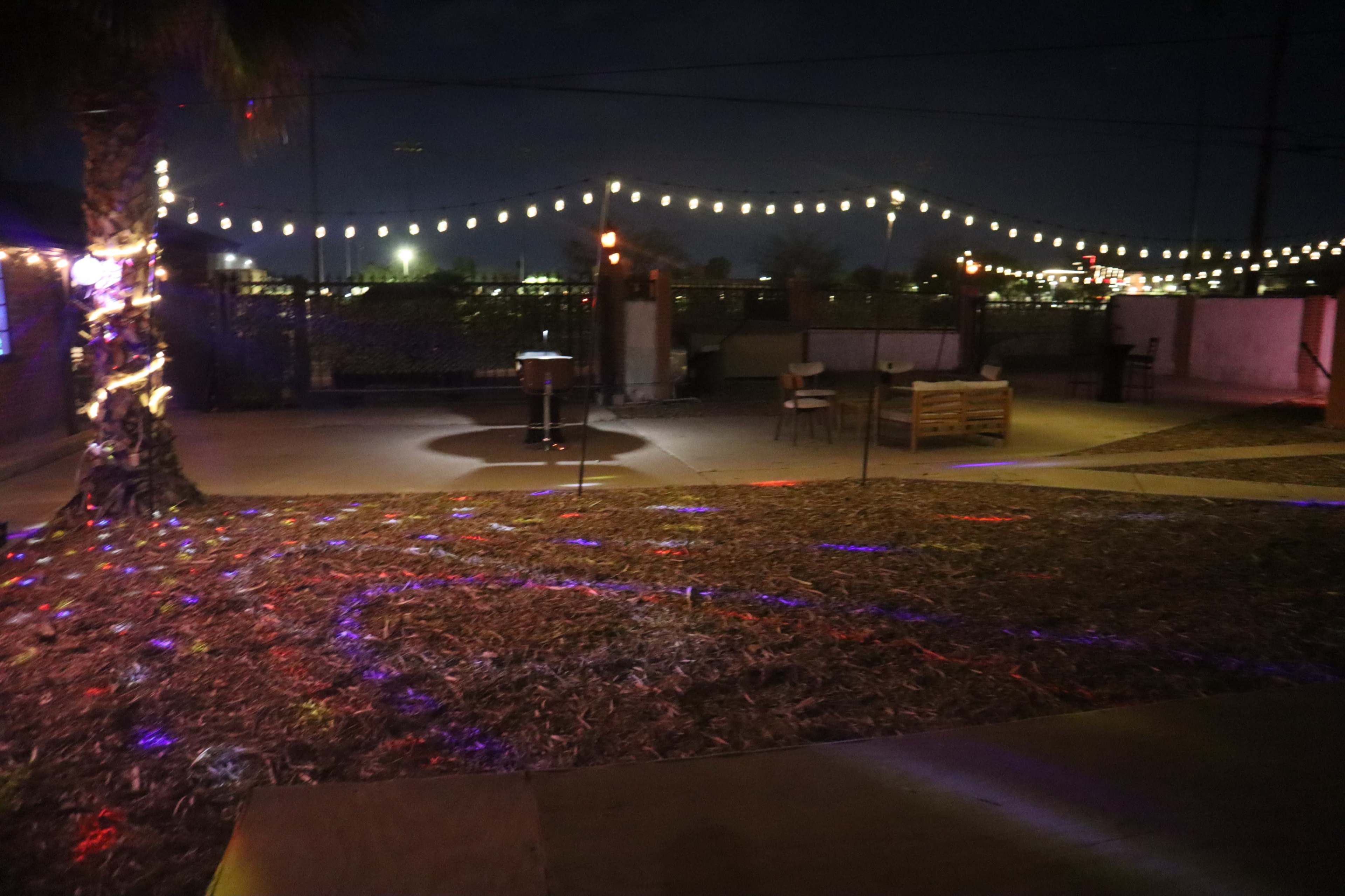 The image shows an outdoor space at night, illuminated by string lights and colored spotlights on the ground, with a few pieces of furniture and a palm tree in the background.