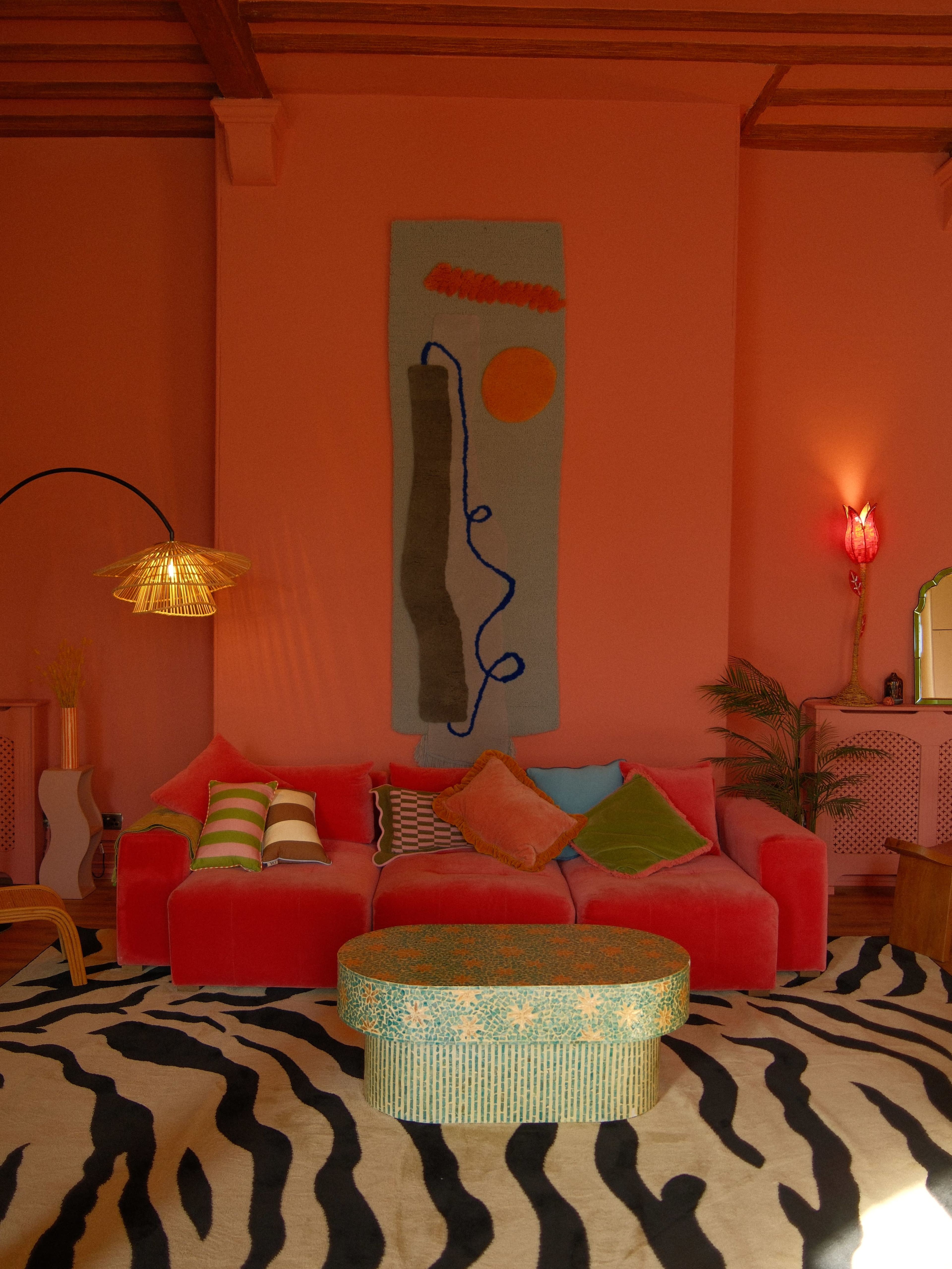 The image shows a vibrant living room with a pink wall, a colorful sectional sofa, a patterned rug, and decorative elements like wall art and lamps.