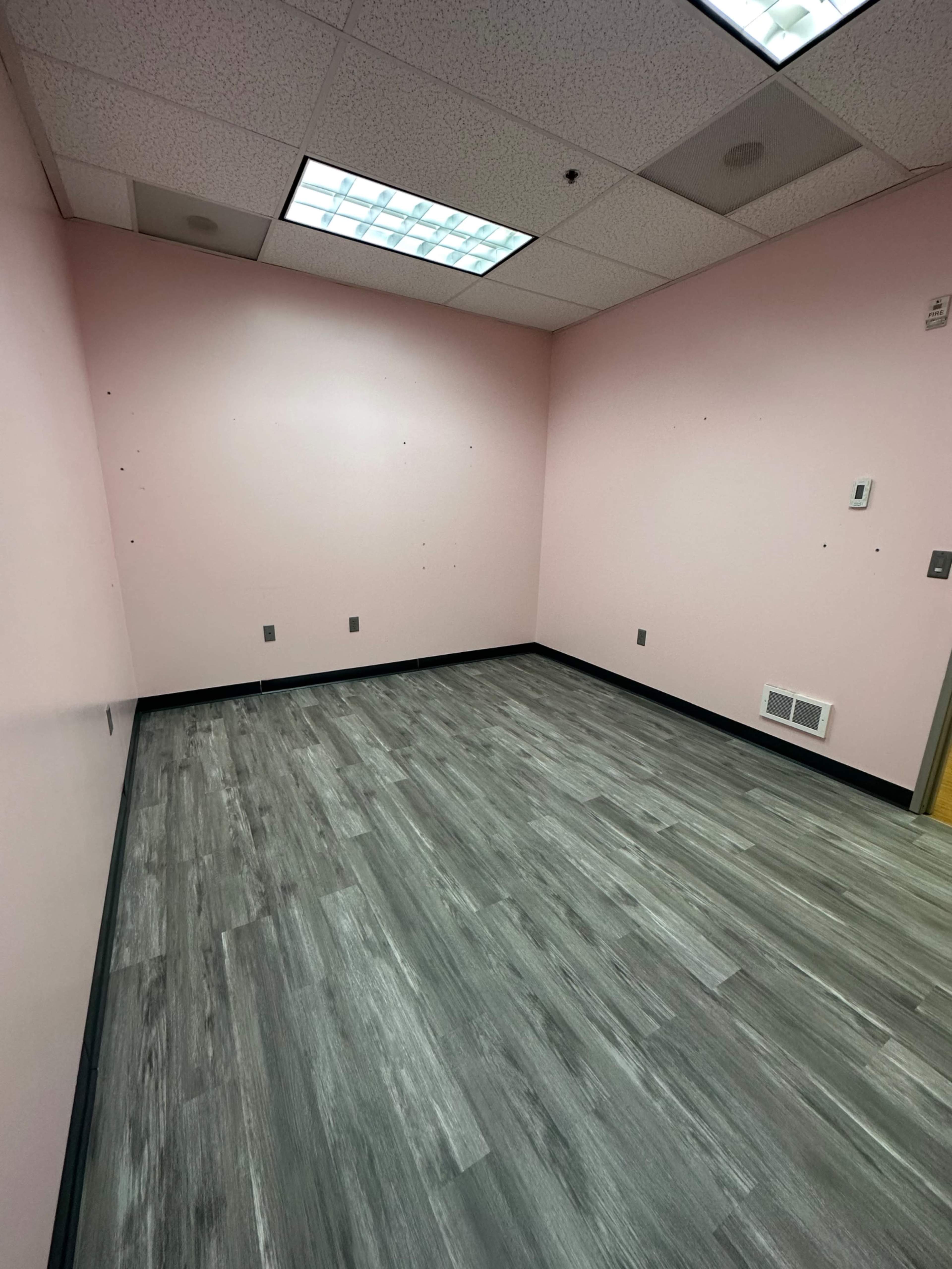 The image shows a vacant room with pink walls and gray laminate flooring, featuring a ceiling with fluorescent lights.