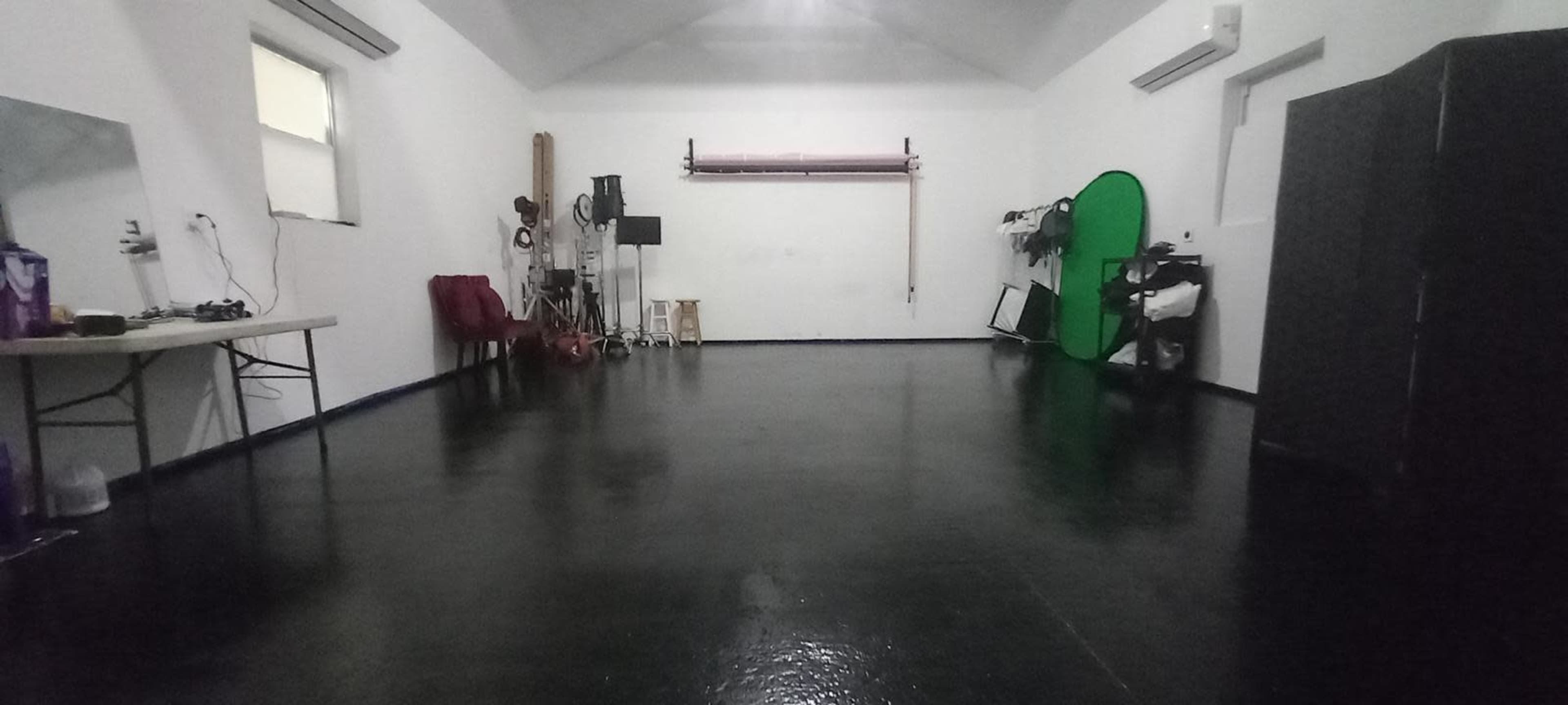 The image shows a spacious, empty room with a polished black floor and various equipment against the walls, including a green screen and a folding table.