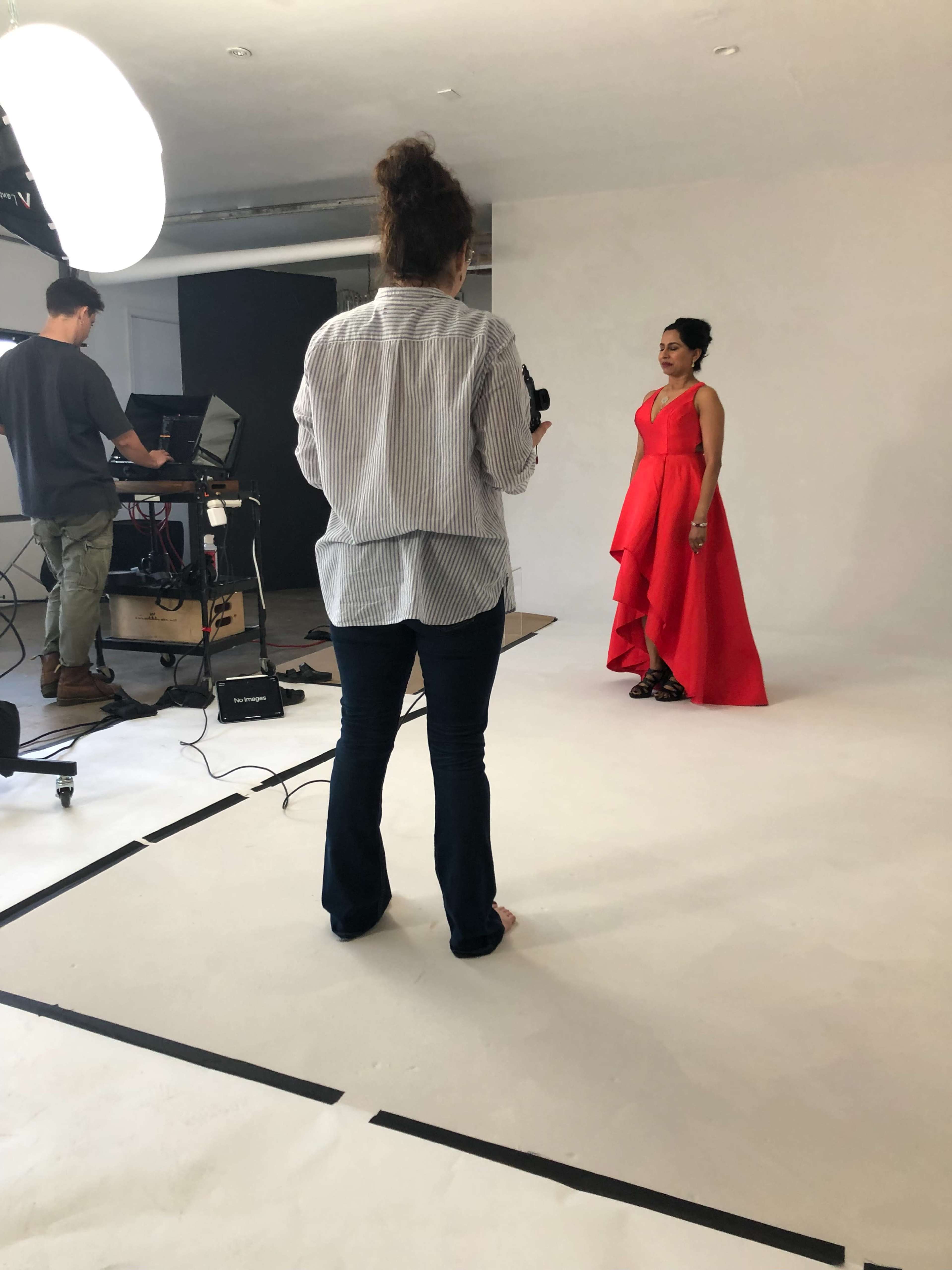A woman in a red dress poses for a photo shoot while a photographer adjusts equipment in the background.