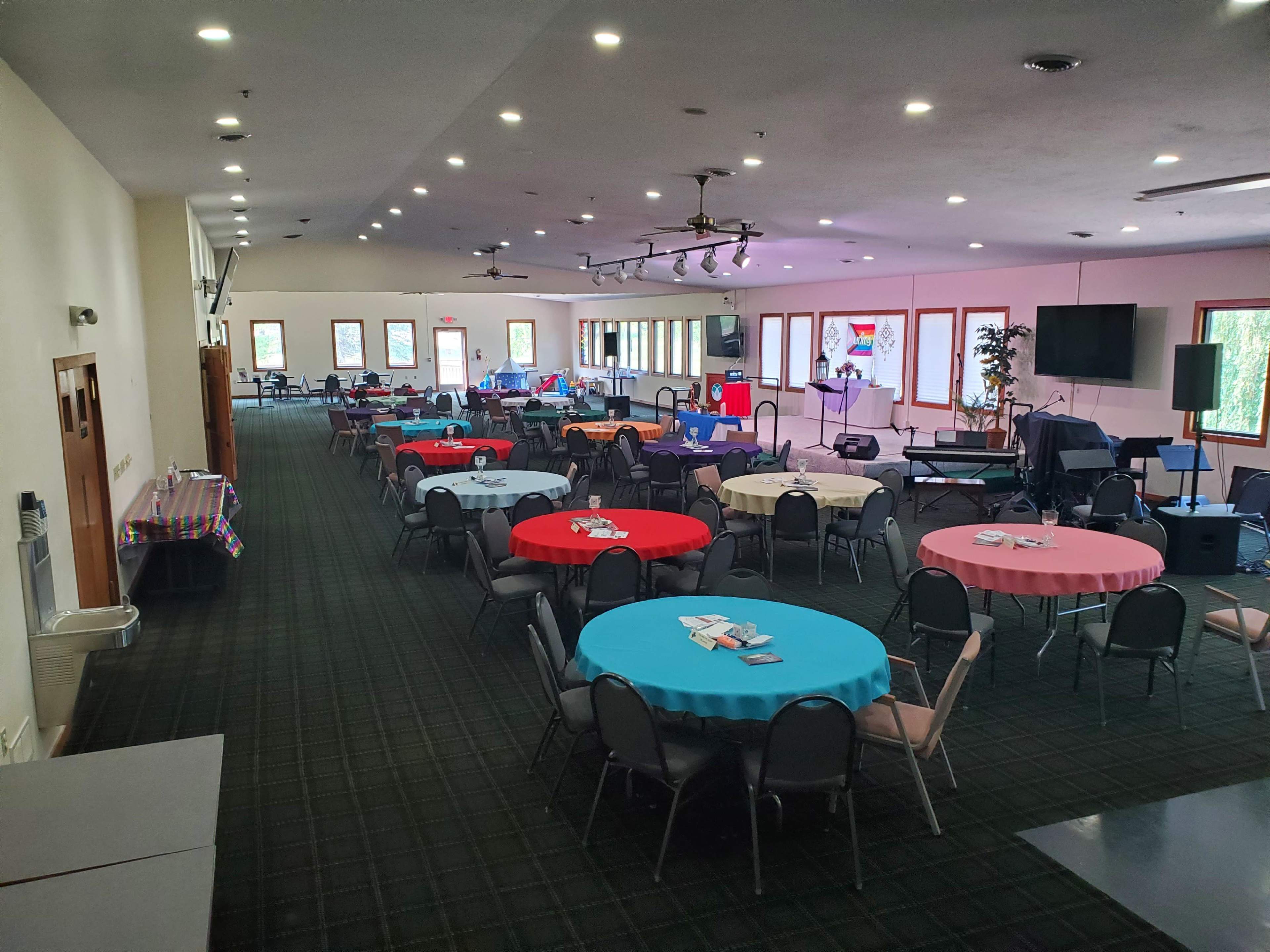 A spacious event hall with several round tables covered in red and blue tablecloths, arranged for a gathering, with a stage and seating area at one end.