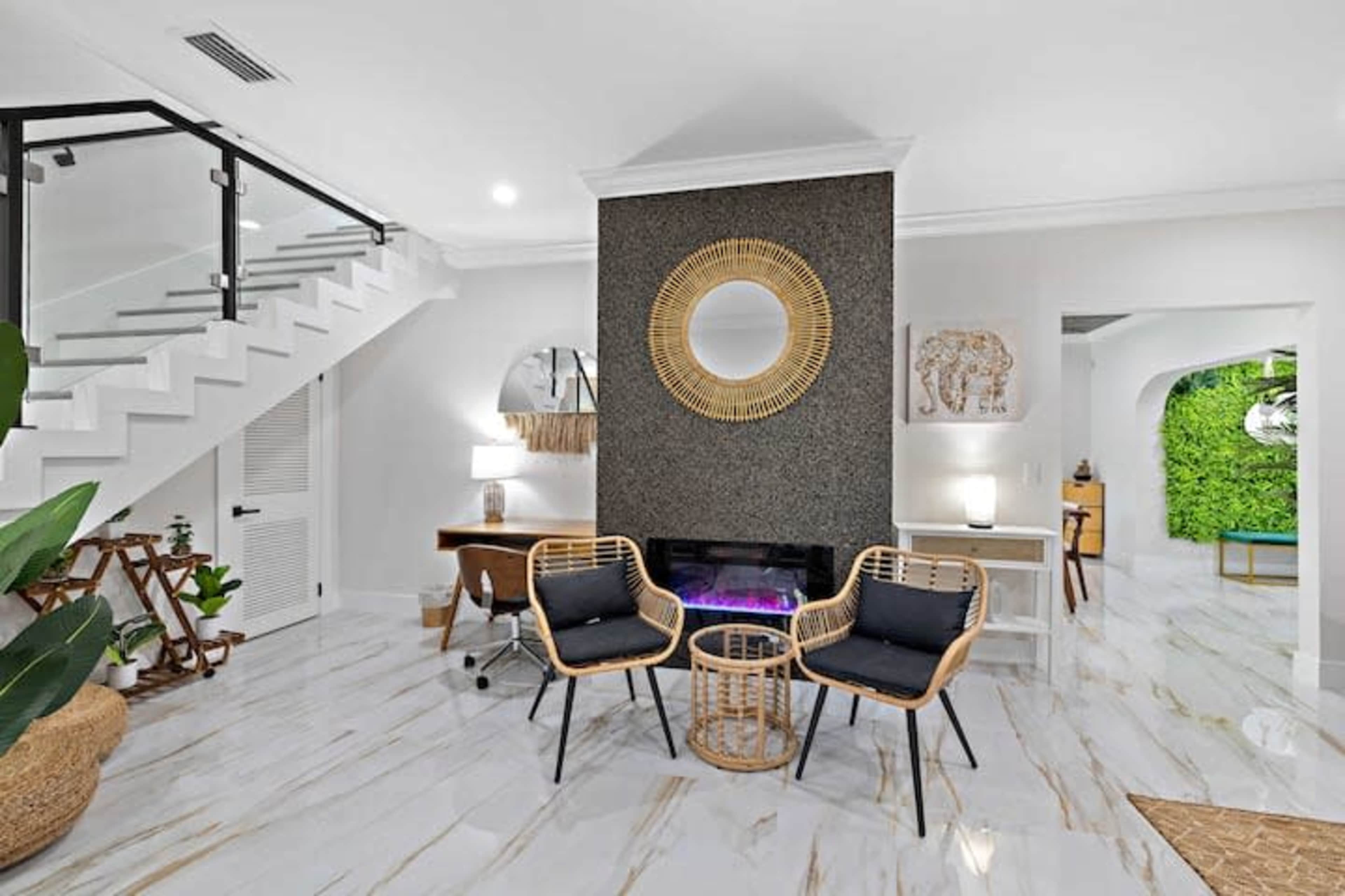 A modern living space featuring a fireplace, two chairs, a round mirror, and a staircase, with greenery visible through an archway.
