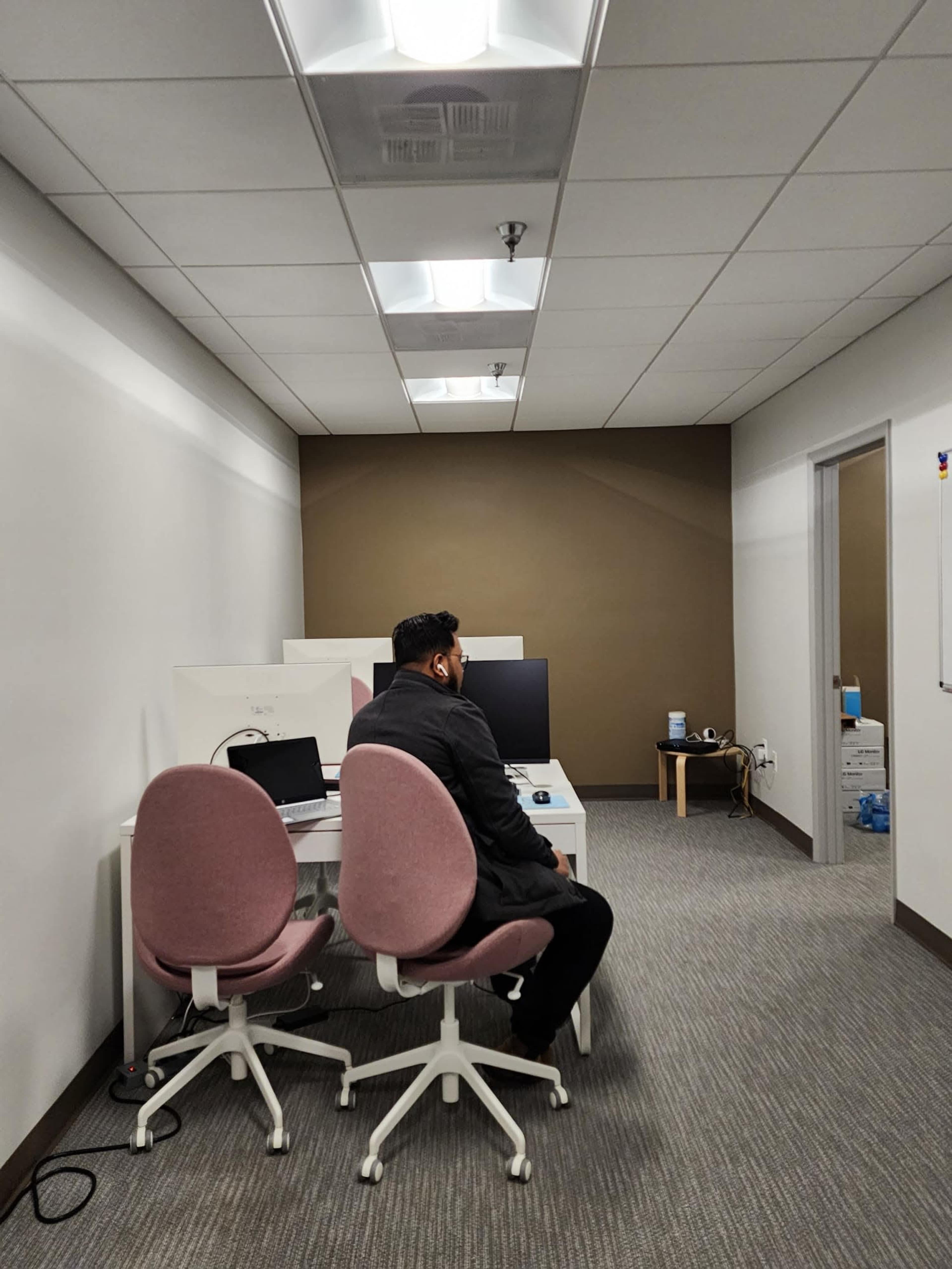 A person sits in an office with two pink chairs facing a desk that holds a computer monitor and a laptop.