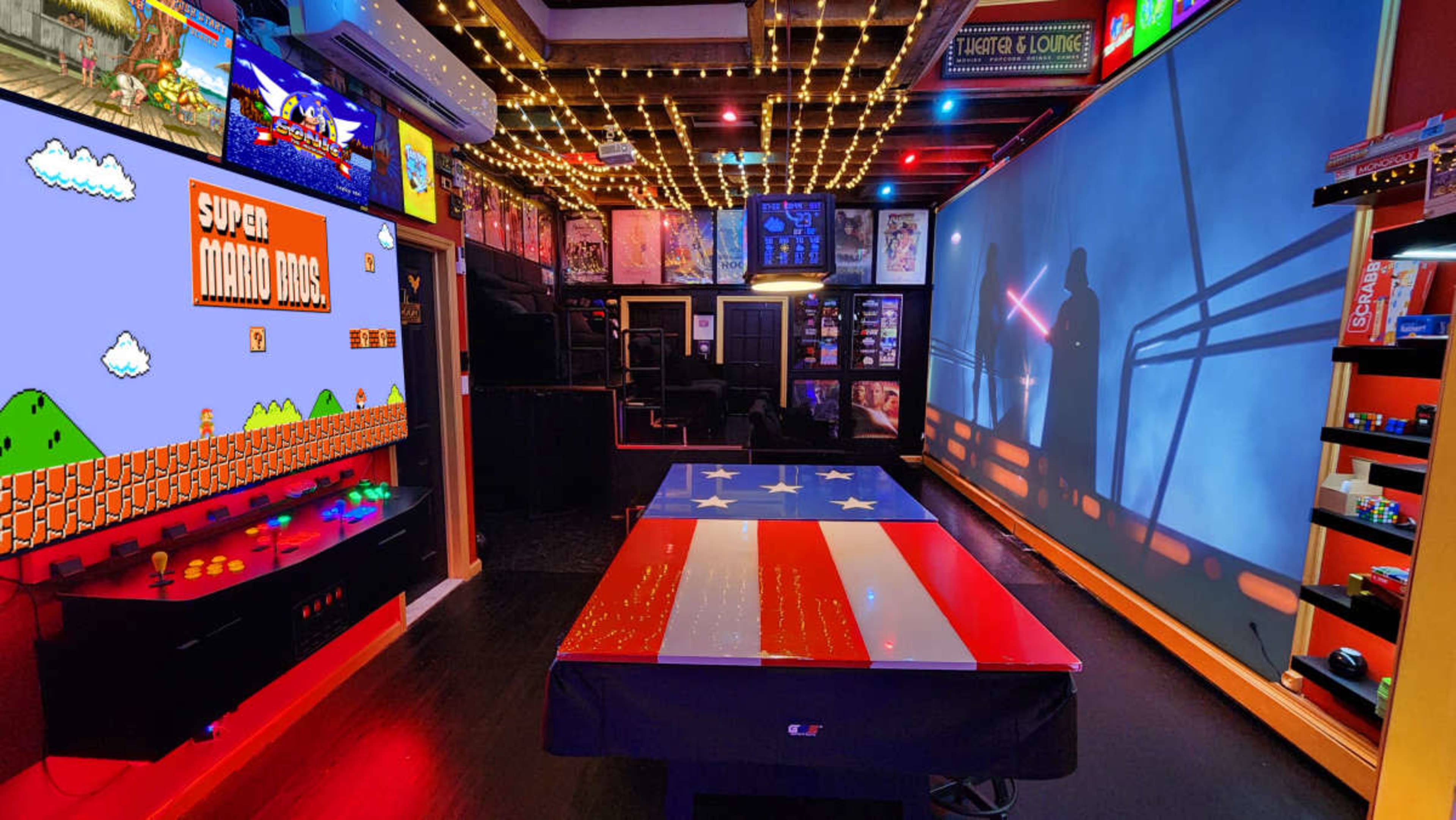 The image features a vibrant game room with a retro arcade setup displaying "Super Mario Bros," a pool table decorated with an American flag, and a large projection on the wall.