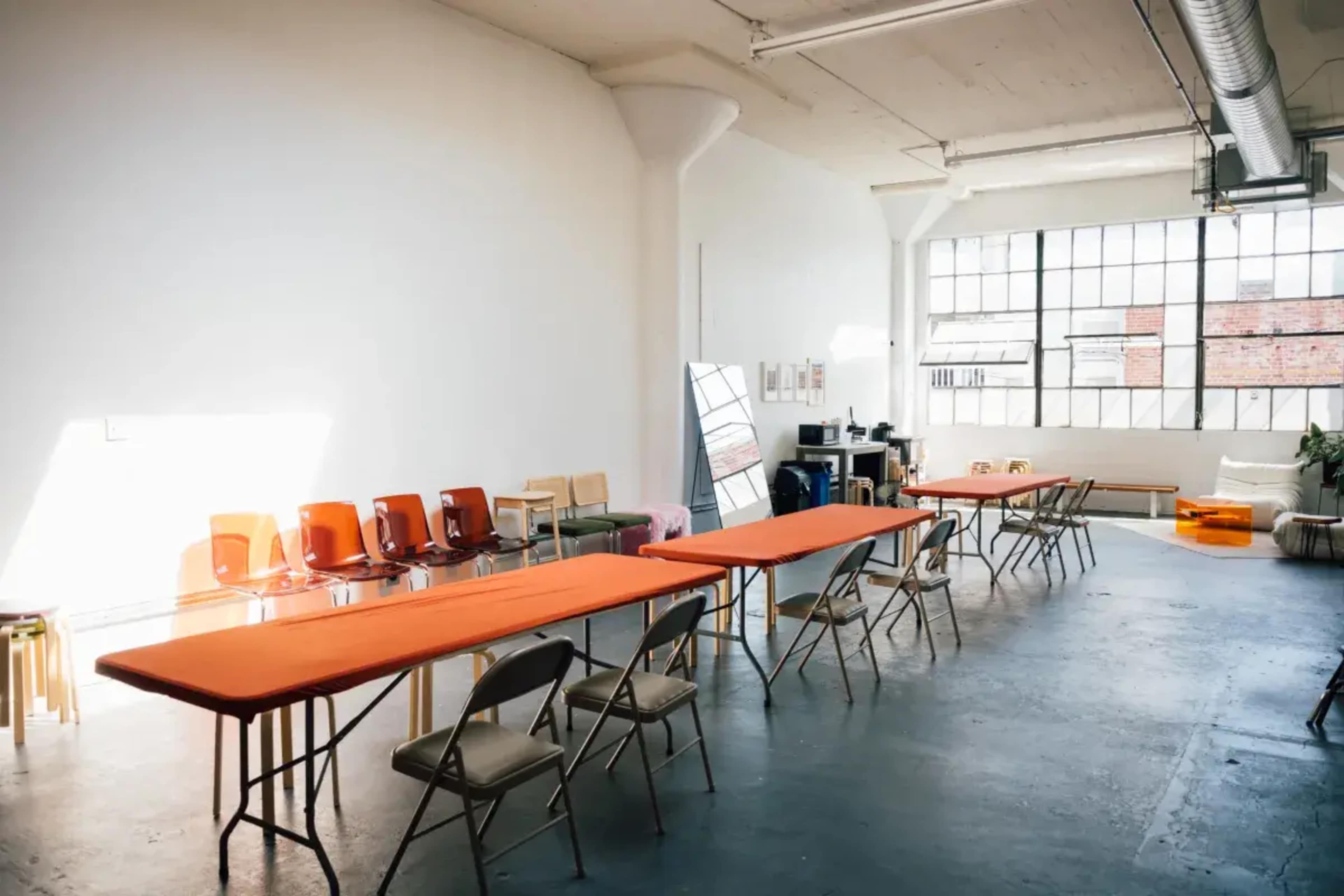 The image shows a spacious, well-lit room with several long orange tables set up, along with metal folding chairs and large windows allowing natural light to enter.