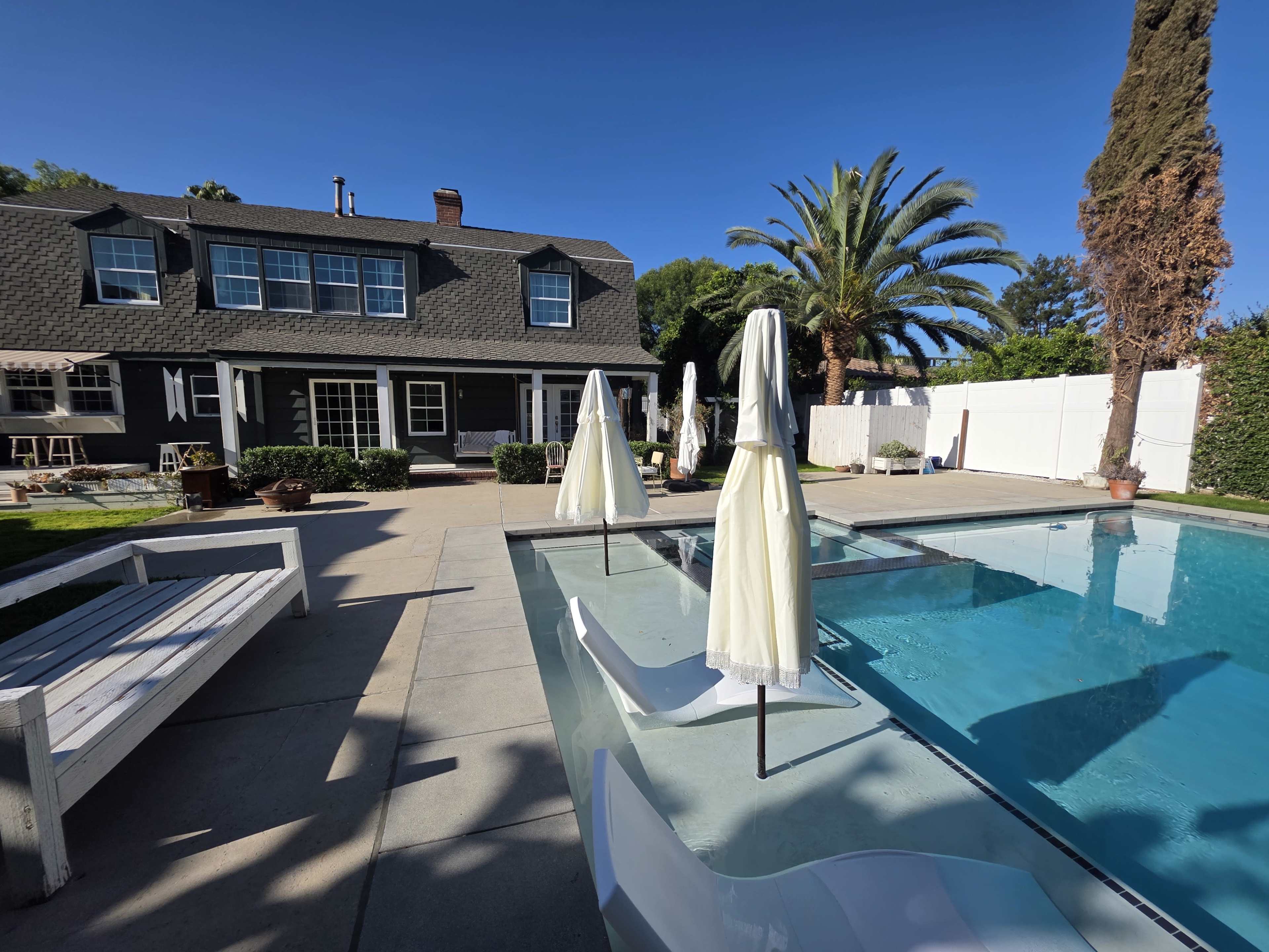 A modern backyard features a swimming pool with white chairs and umbrellas, surrounded by a house and landscaped greenery.