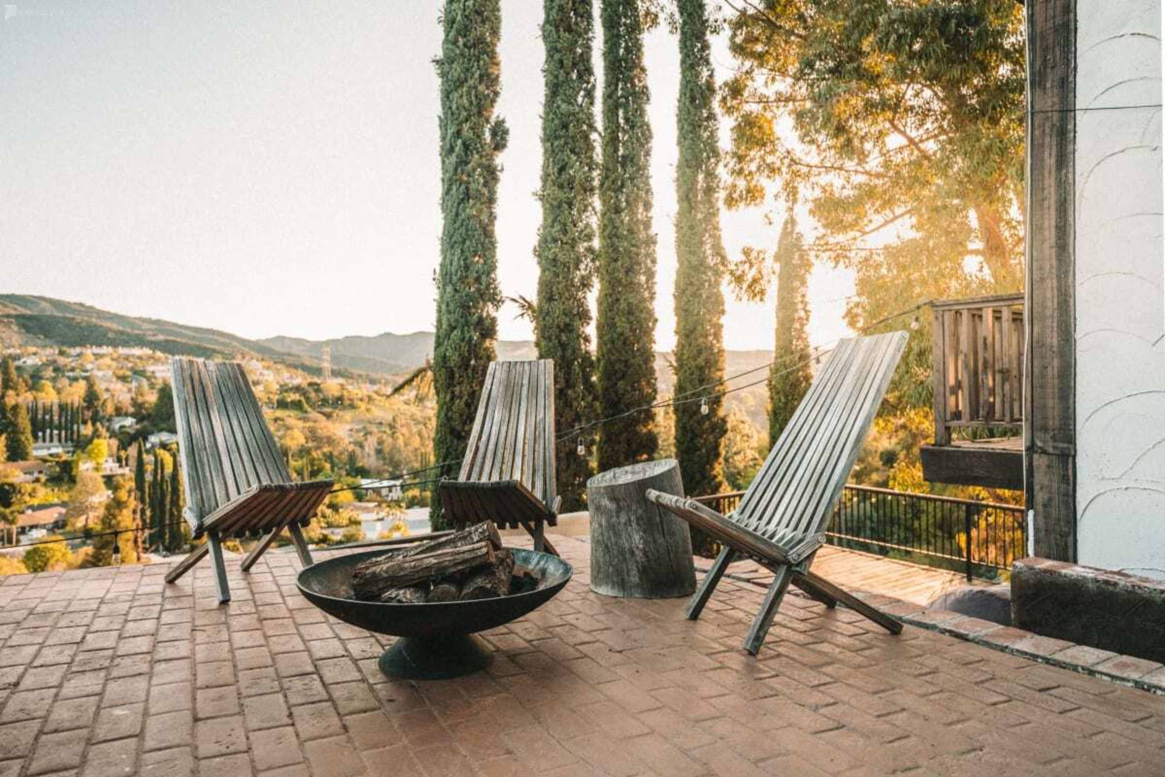 A cozy outdoor seating area features three wooden lounge chairs and a fire pit, set against a backdrop of tall trees and rolling hills at sunset.