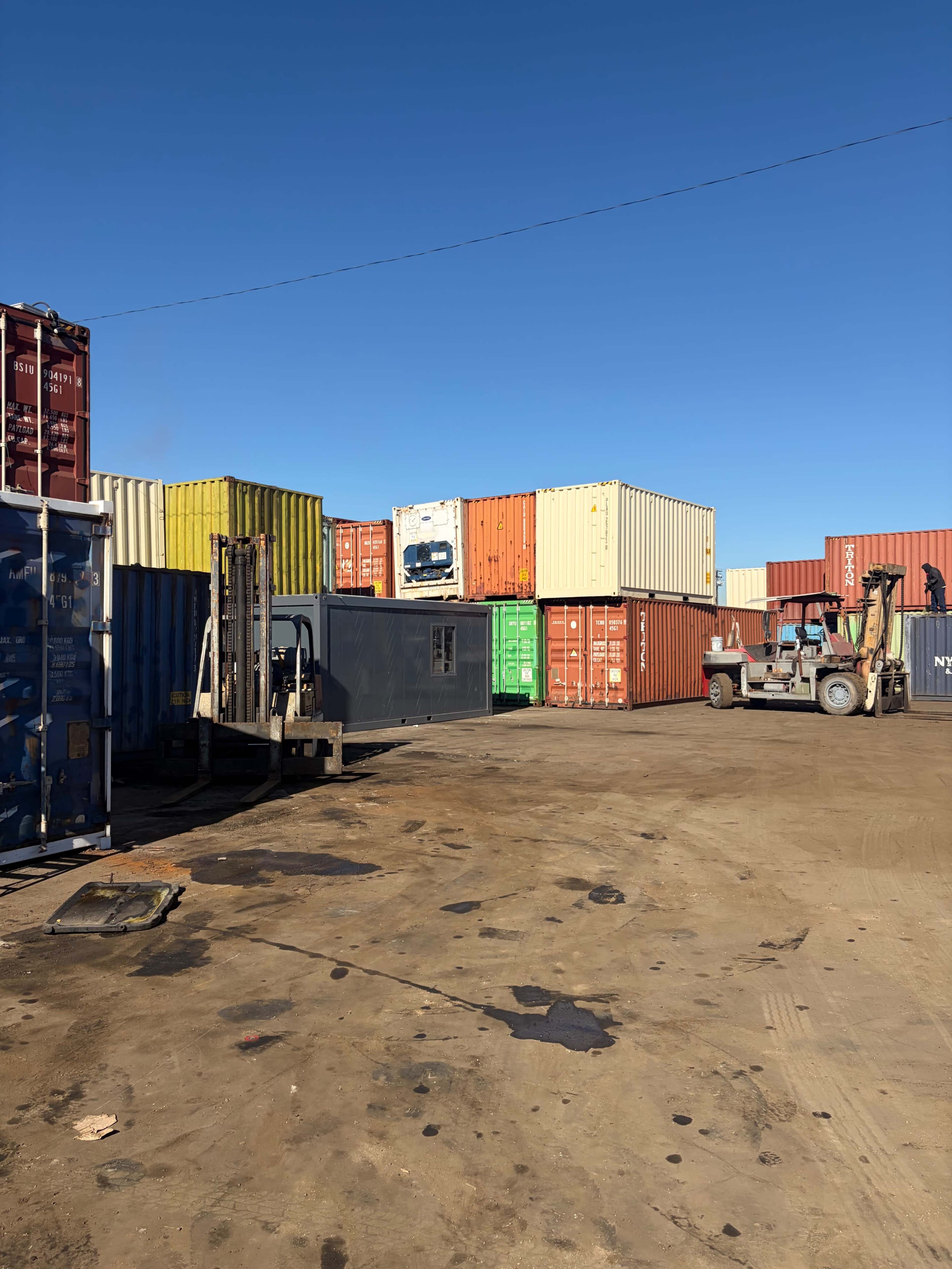 A construction site features stacked shipping containers in various colors, with forklifts and machinery in use on a gravel surface under a clear blue sky.