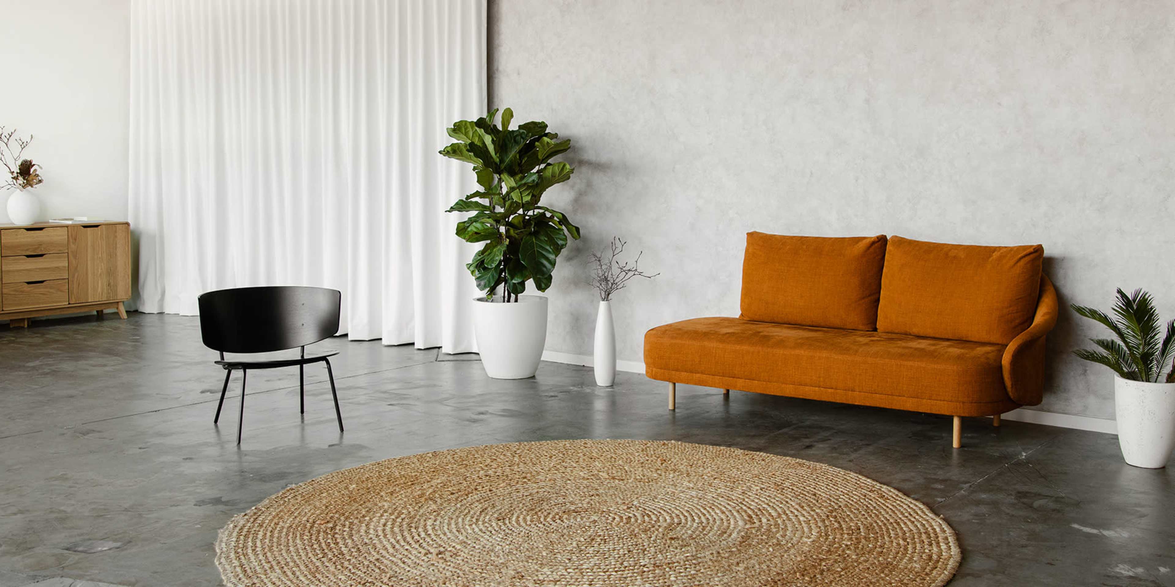 A modern living space featuring an orange sofa, a black chair, a round jute rug, and large potted plants against a light gray wall.