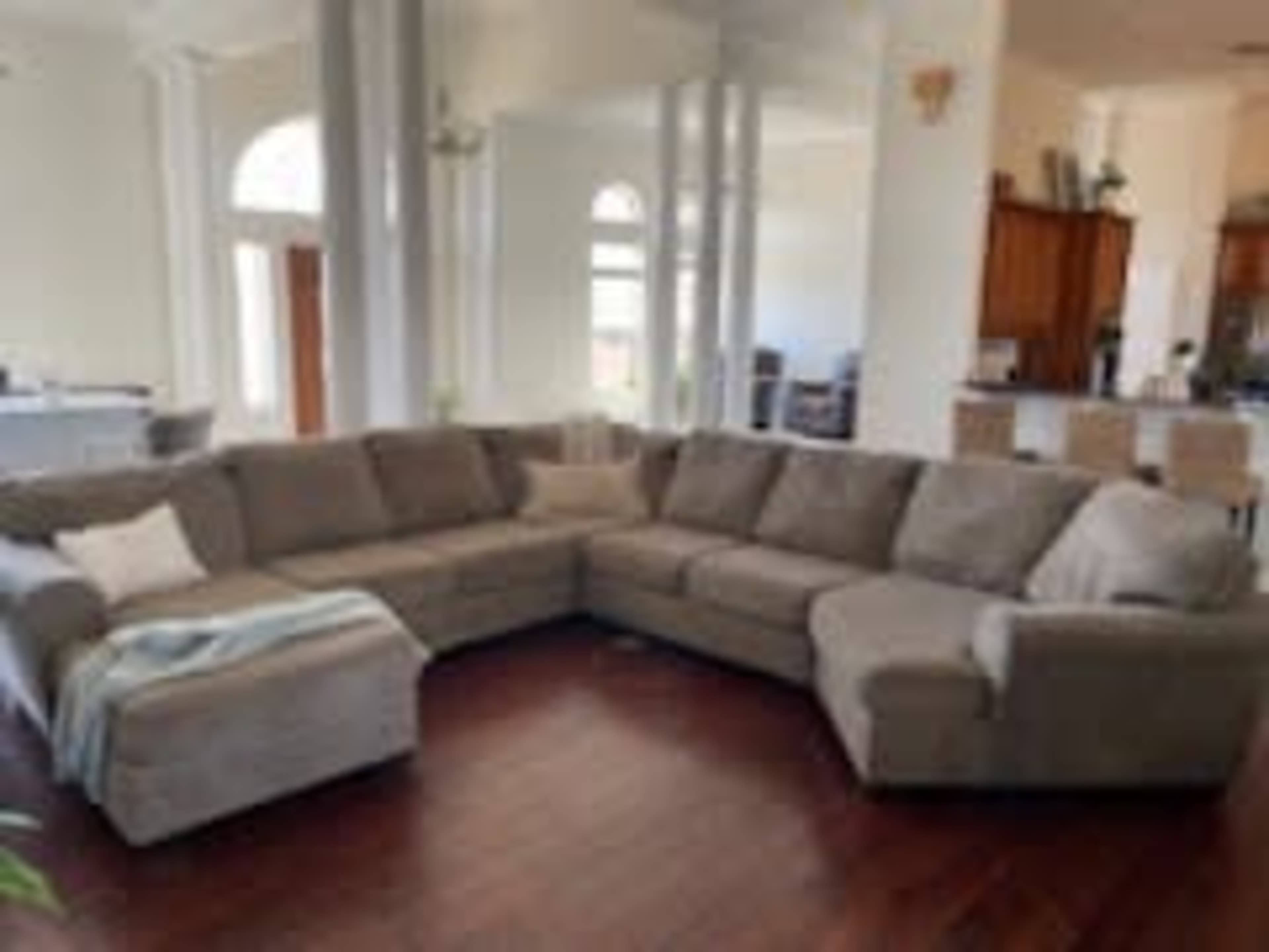 A large, sectional sofa occupies a bright, open living space with wooden flooring and a view of a kitchen in the background.