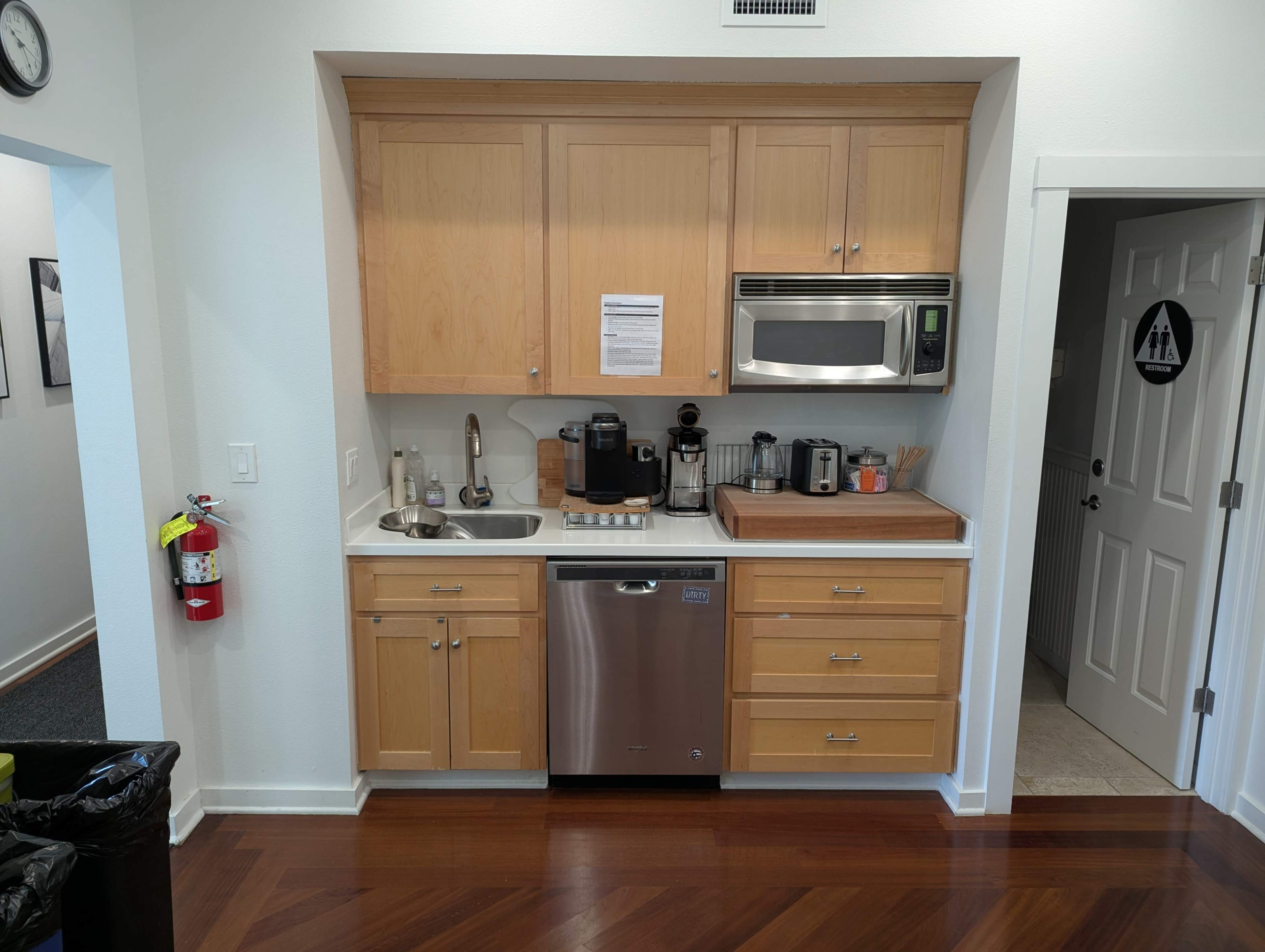 The image shows a kitchen area with wooden cabinetry, a sink, a dishwasher, a microwave, and various small appliances neatly arranged on the counter.