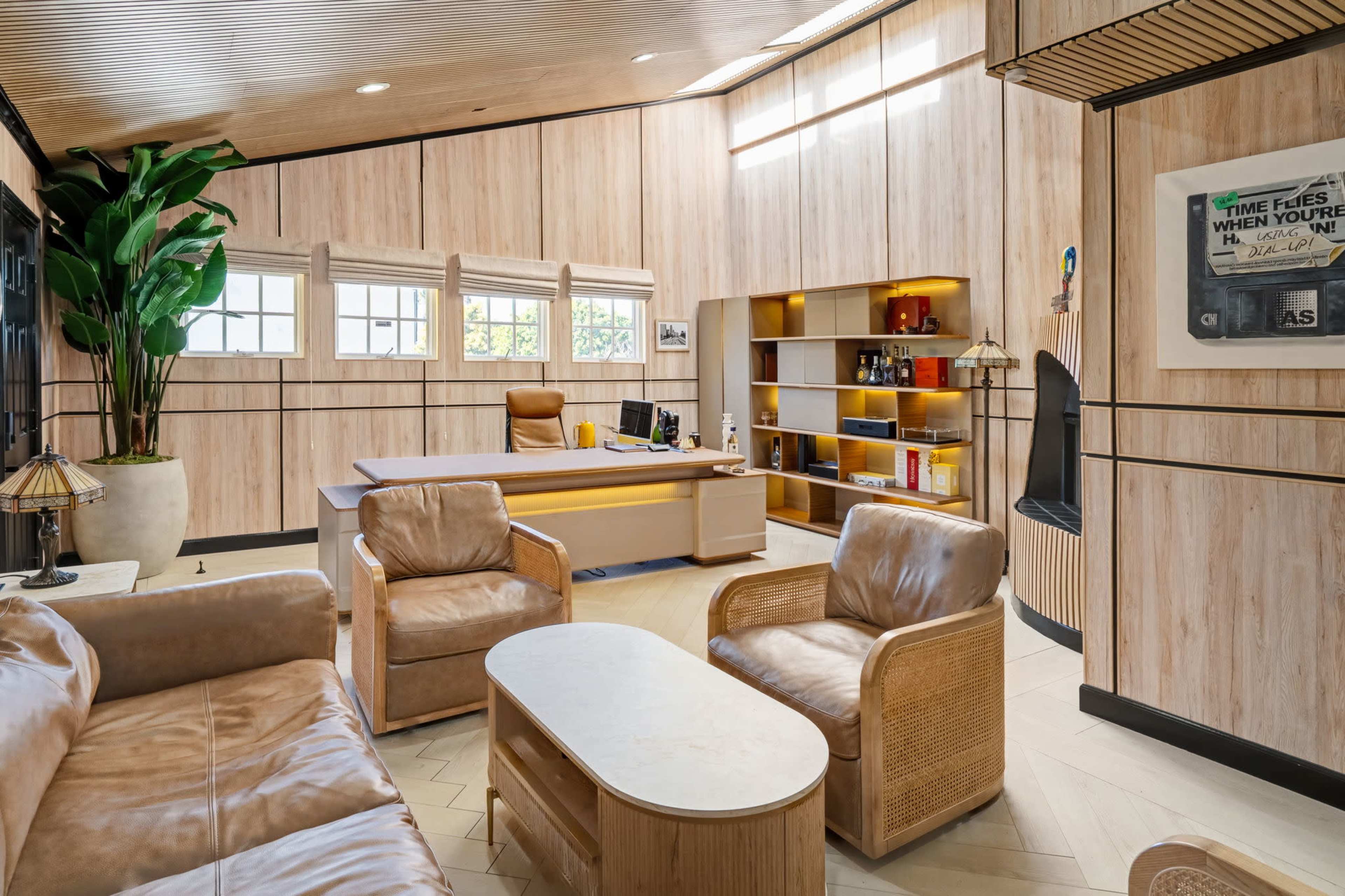 The image shows a modern office interior with wooden paneling, a desk, bookshelves, and seating arrangements.