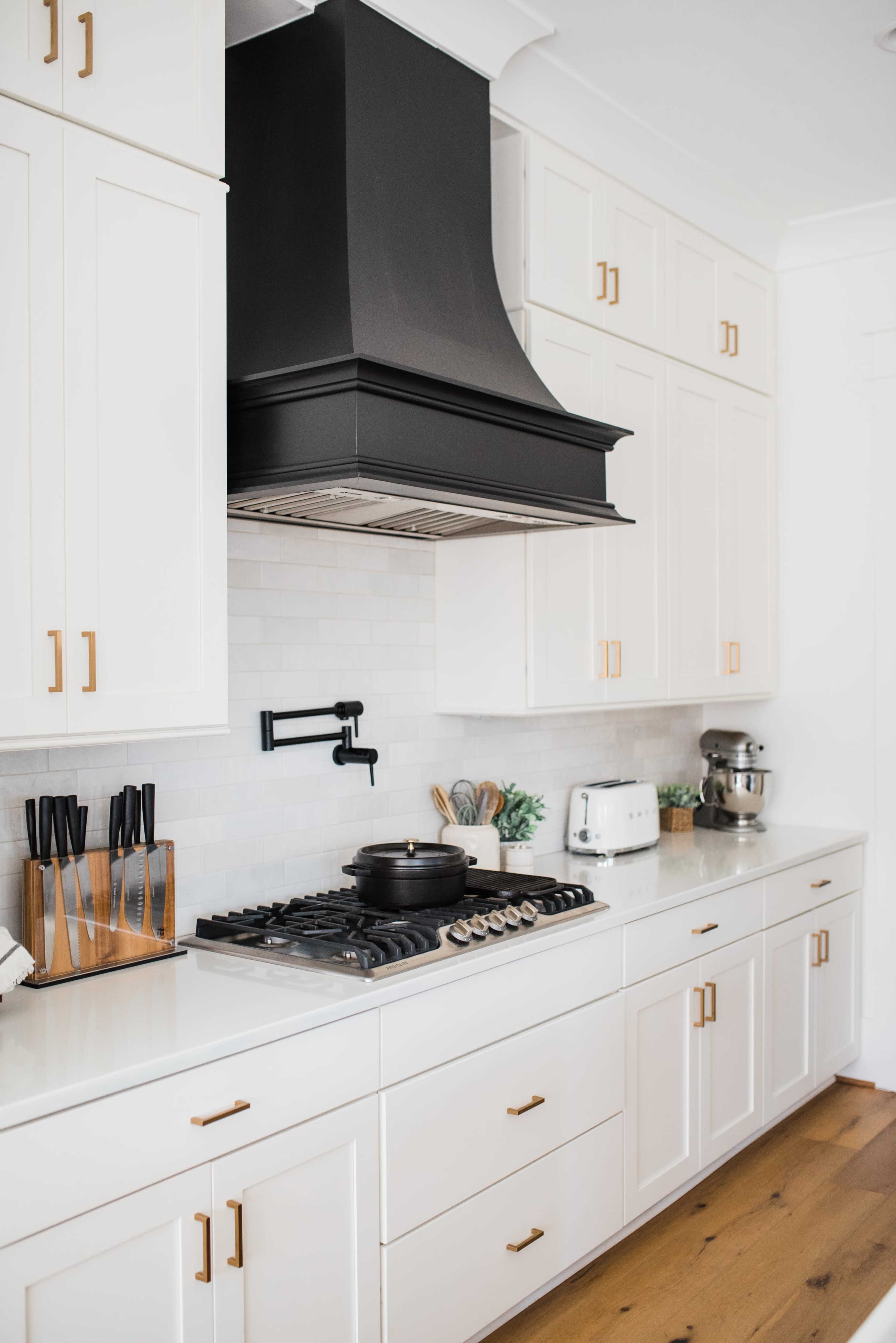 A modern kitchen featuring white cabinetry, a black range hood, and stainless steel appliances, with a gas stovetop and wooden flooring.