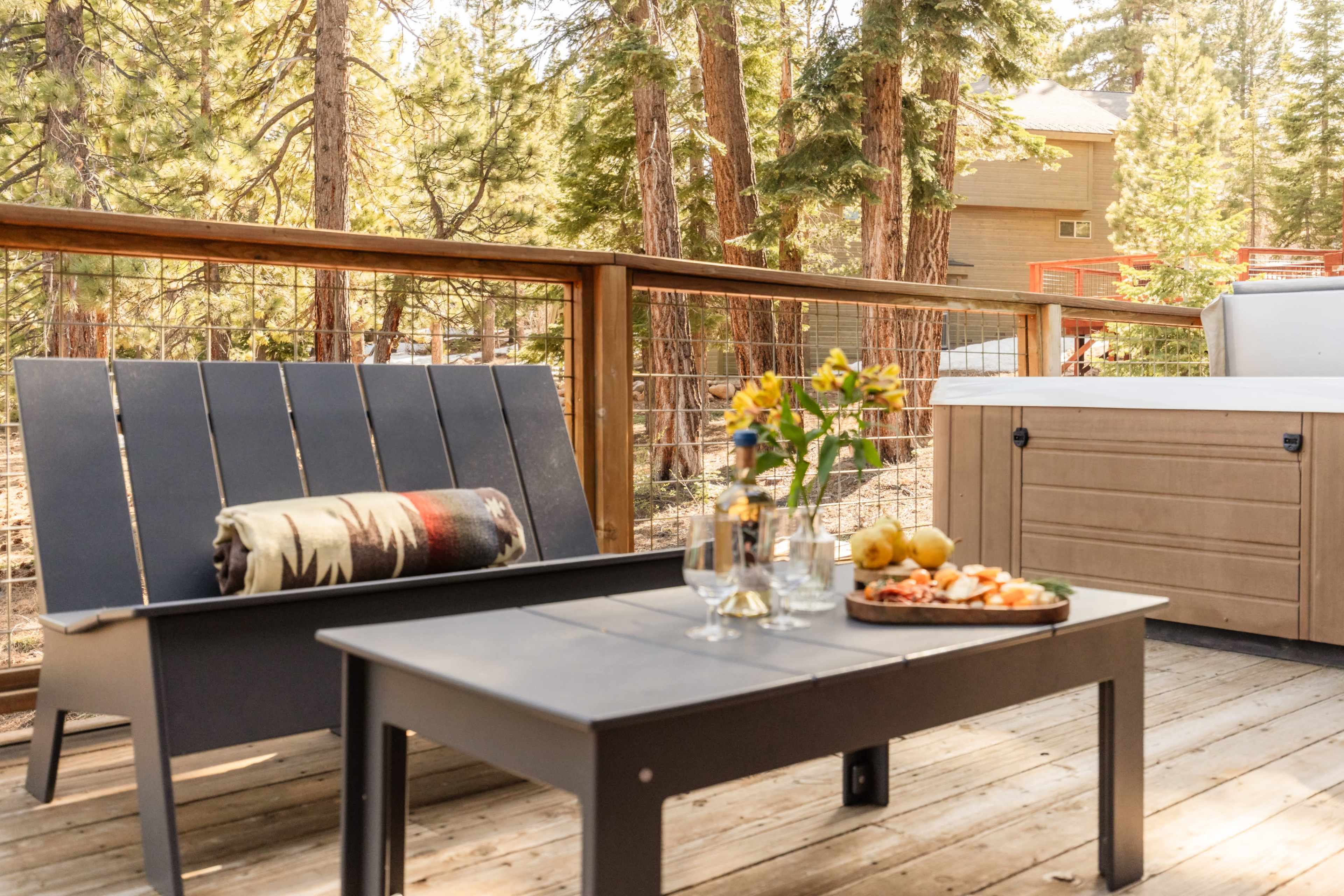 A wooden deck features a gray lounge chair, a coffee table with a plate of snacks, and a flower vase, surrounded by tall trees and a distant building.
