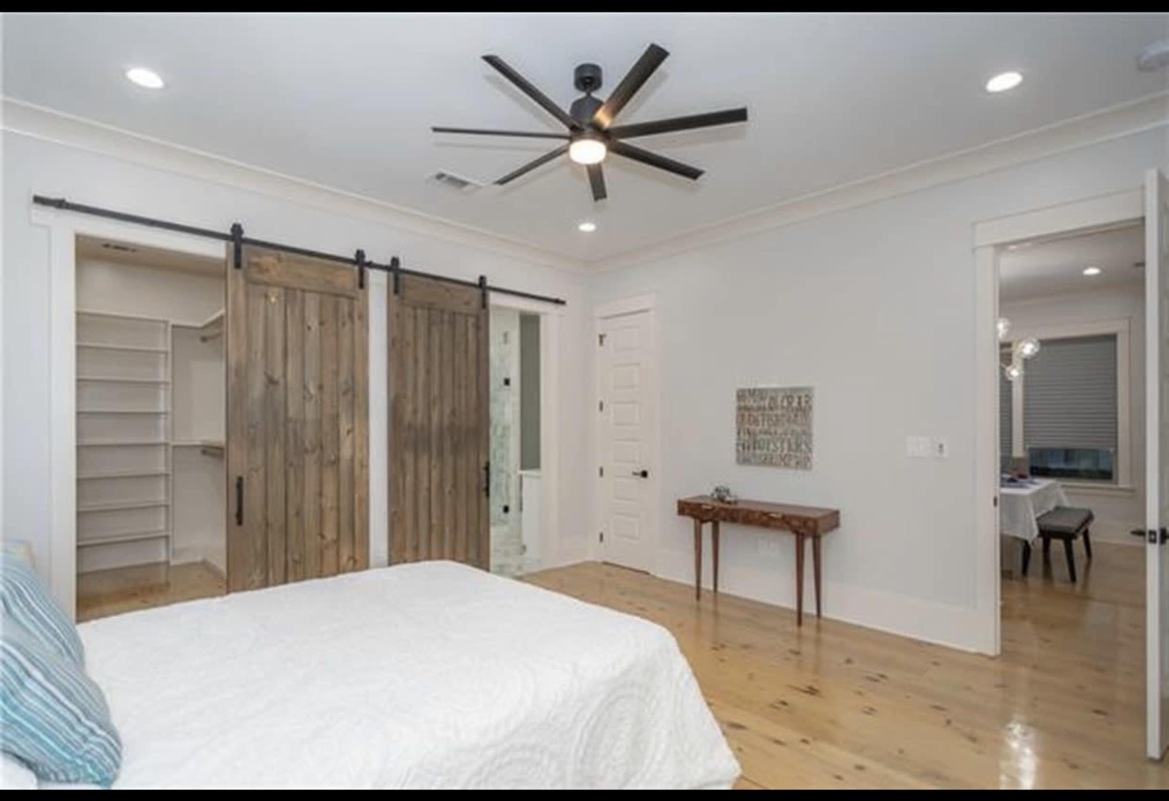 The image depicts a modern bedroom featuring a white bedspread, sliding wooden barn doors, and a small wooden table against the wall.