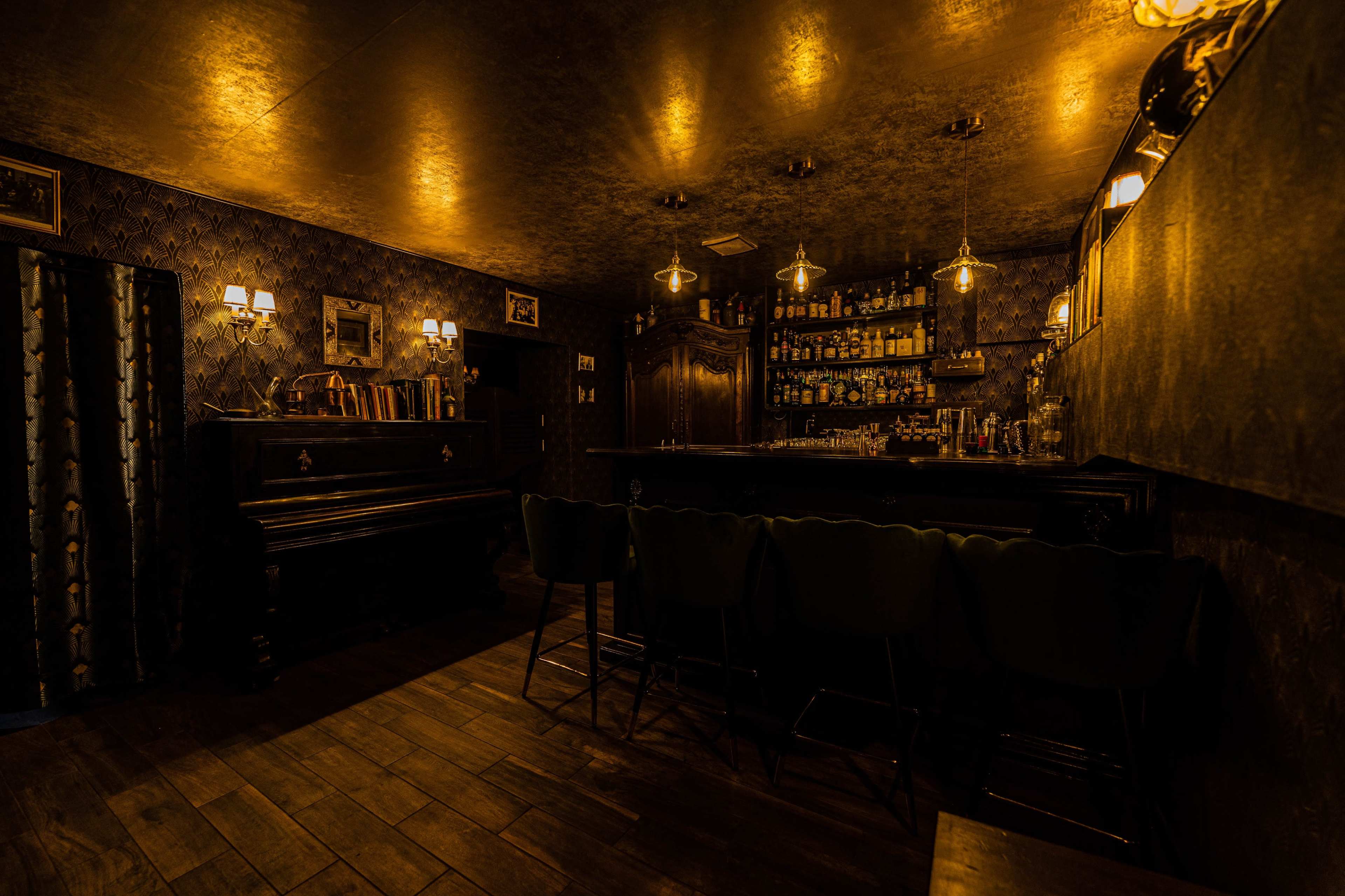 A dimly lit bar features a piano and a well-stocked backlit shelf of liquor against a patterned wall.