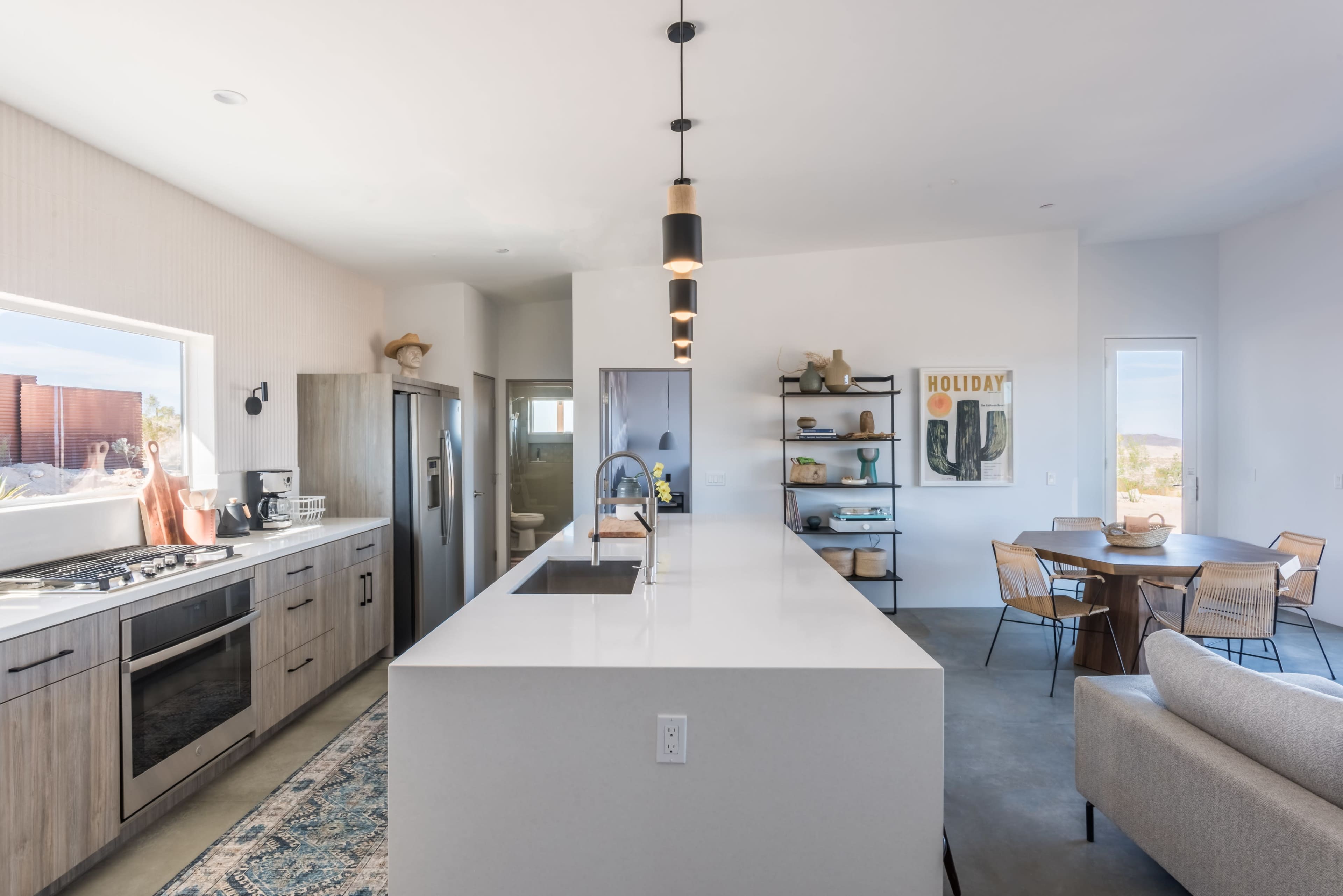 The image shows a modern kitchen with a large island, stainless steel appliances, and a dining area featuring a round table and chairs.