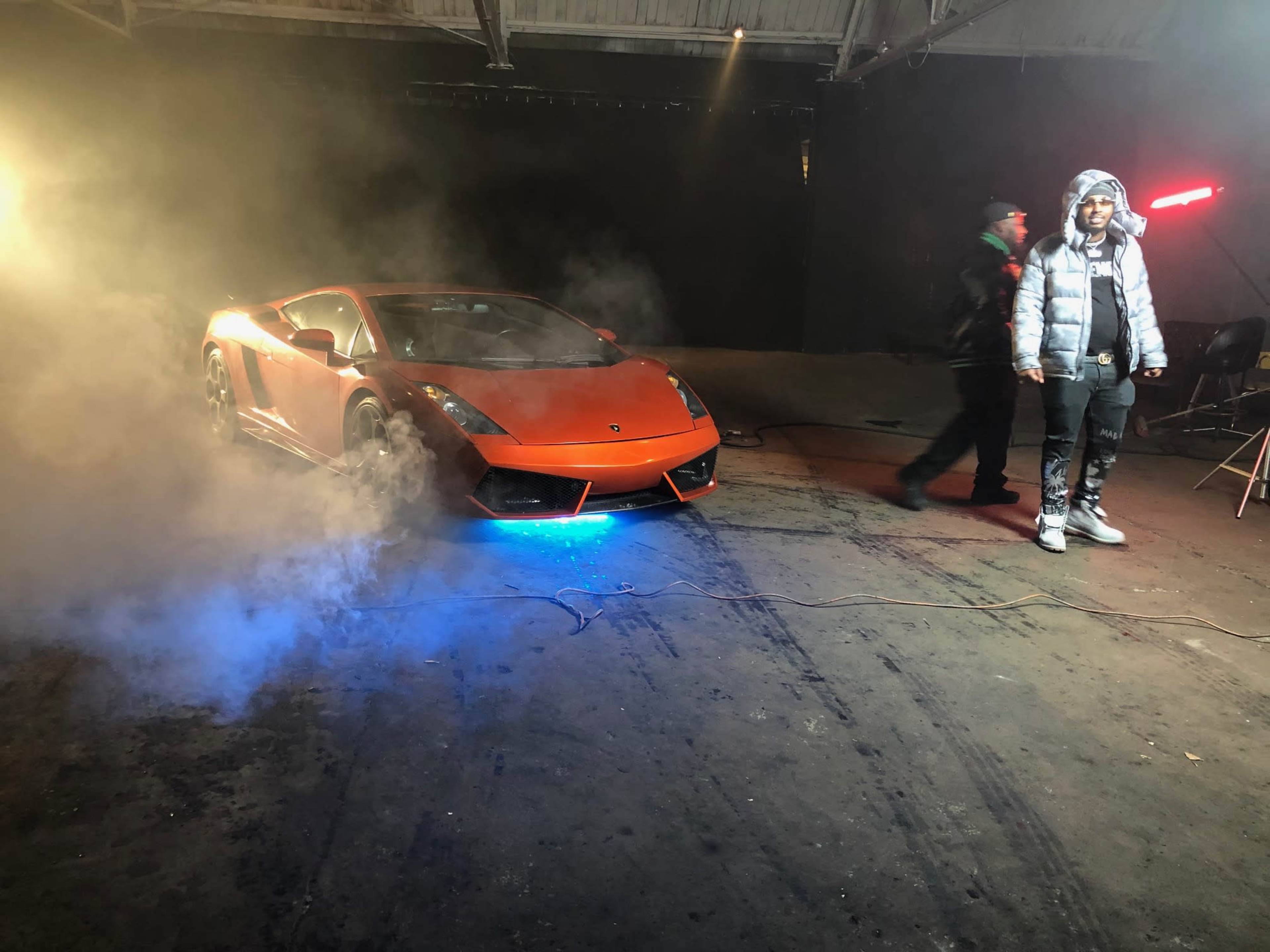 A bright orange Lamborghini Gallardo is parked in a dimly lit space with fog and a figure standing nearby.