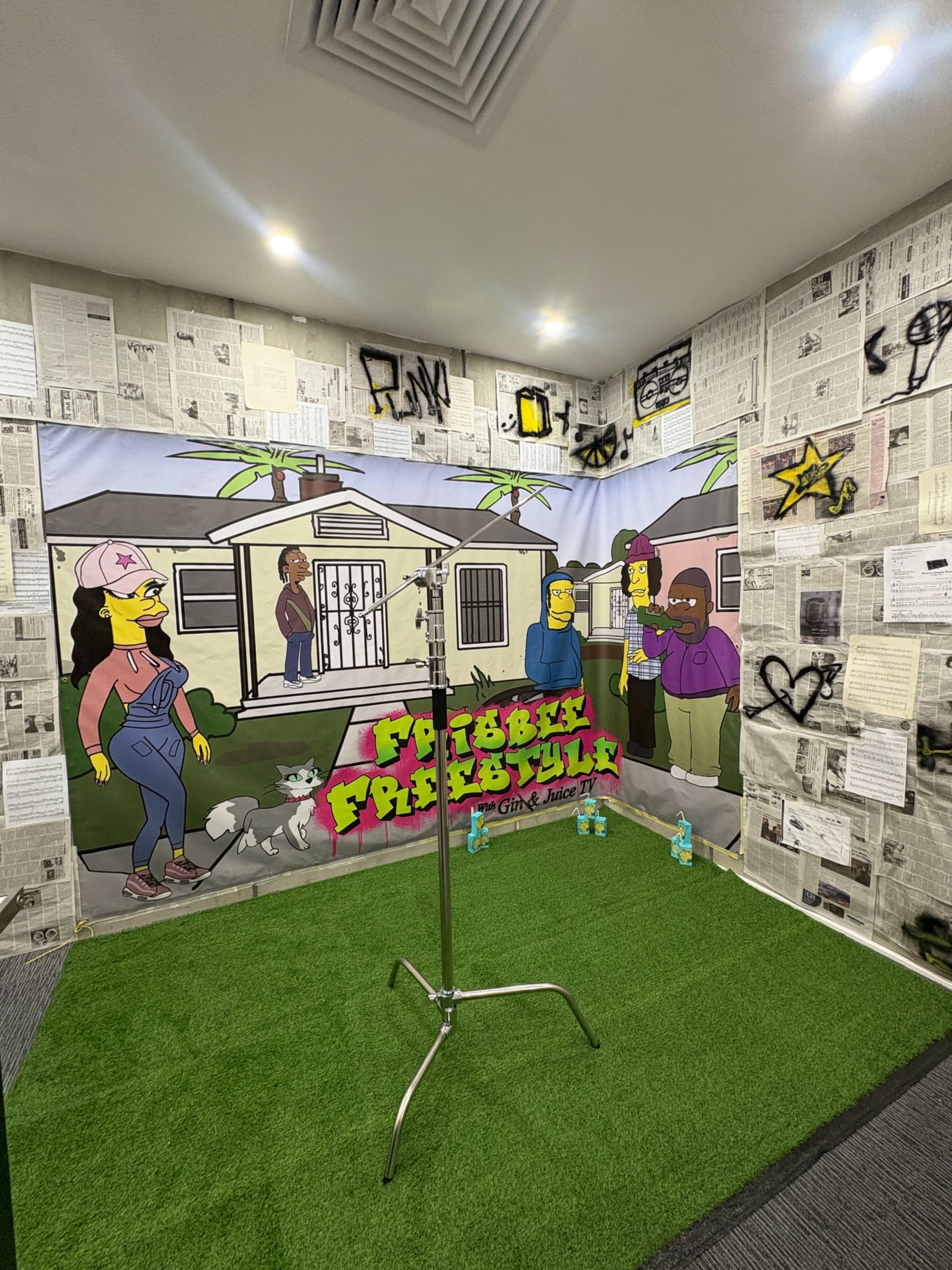 The image shows a themed backdrop featuring cartoon characters from "The Simpsons," set against a wall covered in newspaper clippings and graffiti, with artificial grass on the floor.