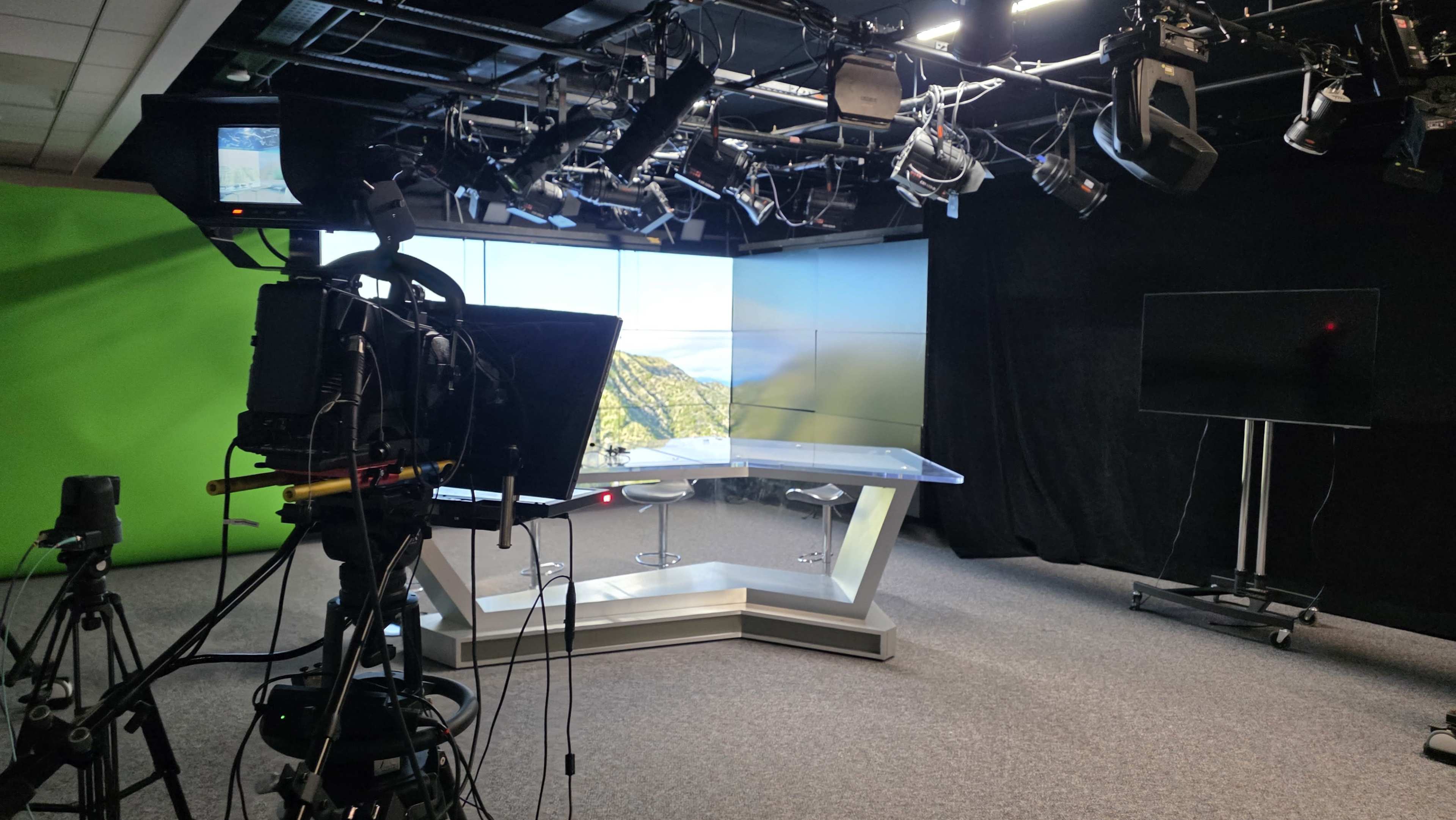 The image shows a television studio setup with a clear glass news desk, two chairs, and various cameras positioned in front of a green screen and a large monitor displaying a scenic backdrop.