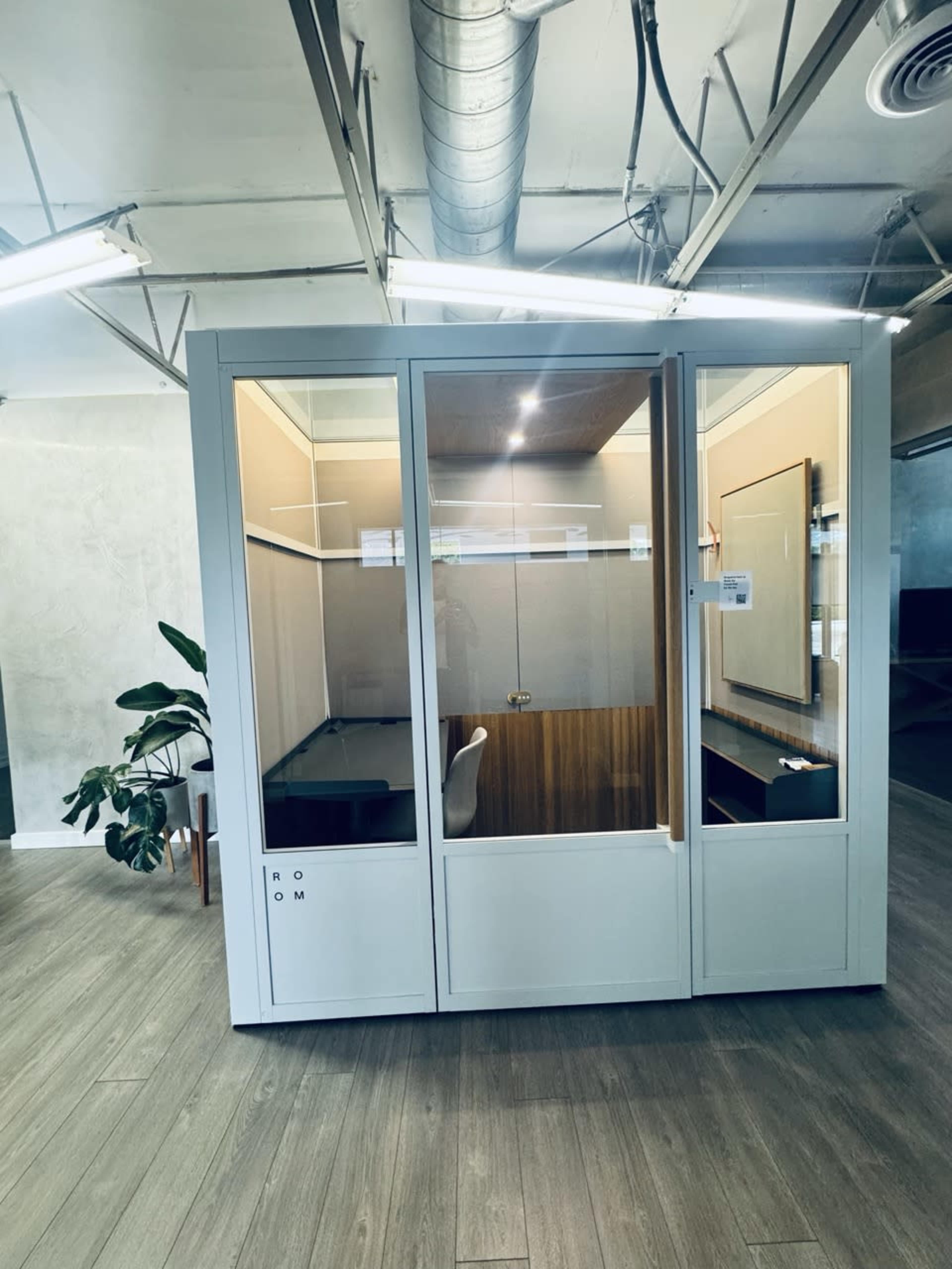 The image shows a modern glass-enclosed workspace with a small desk and a chair, surrounded by a minimalistic interior design.