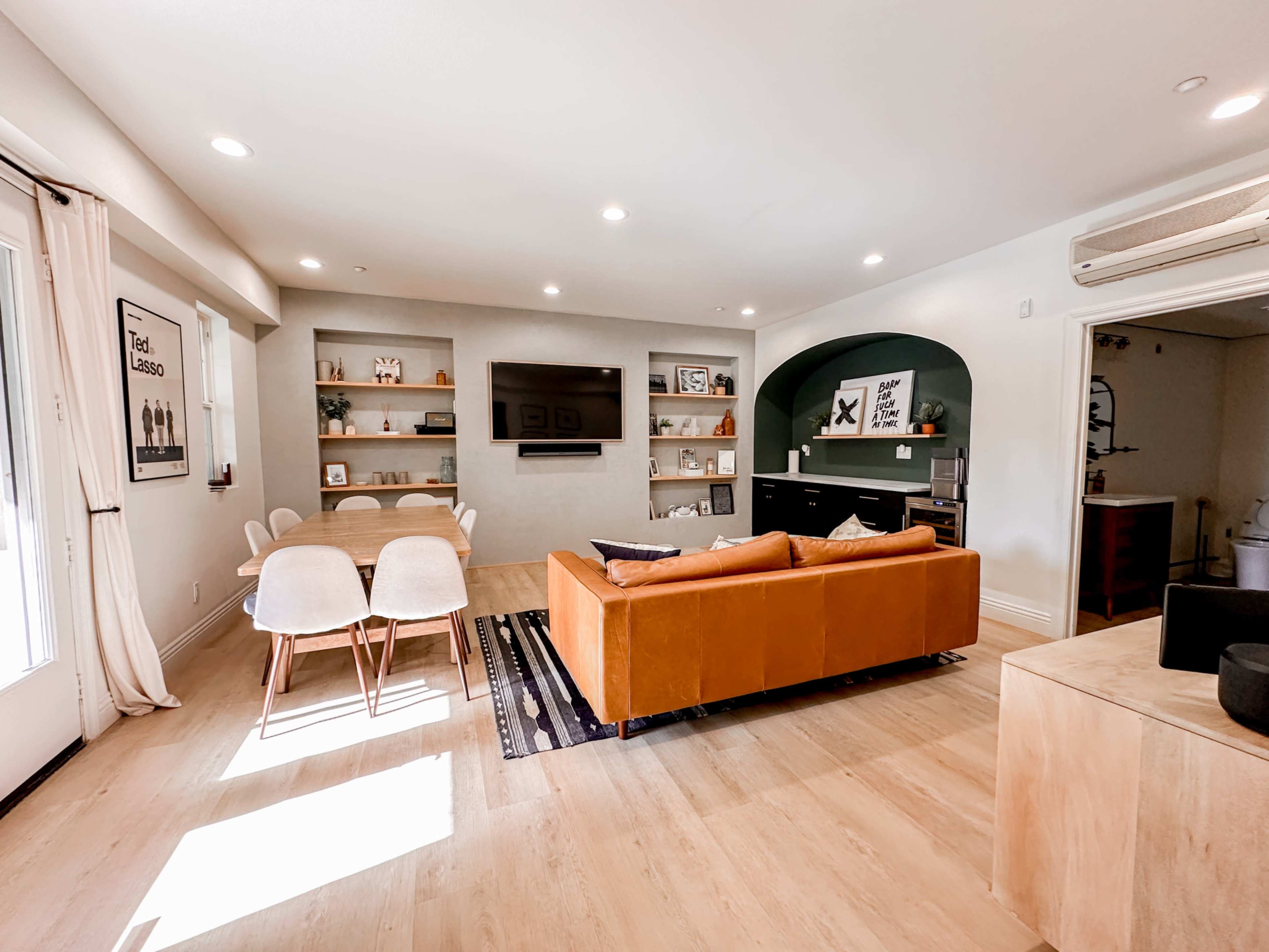 A modern living room with a brown couch, a dining area with white chairs, a flat-screen TV mounted on the wall, and shelves displaying various decorations.