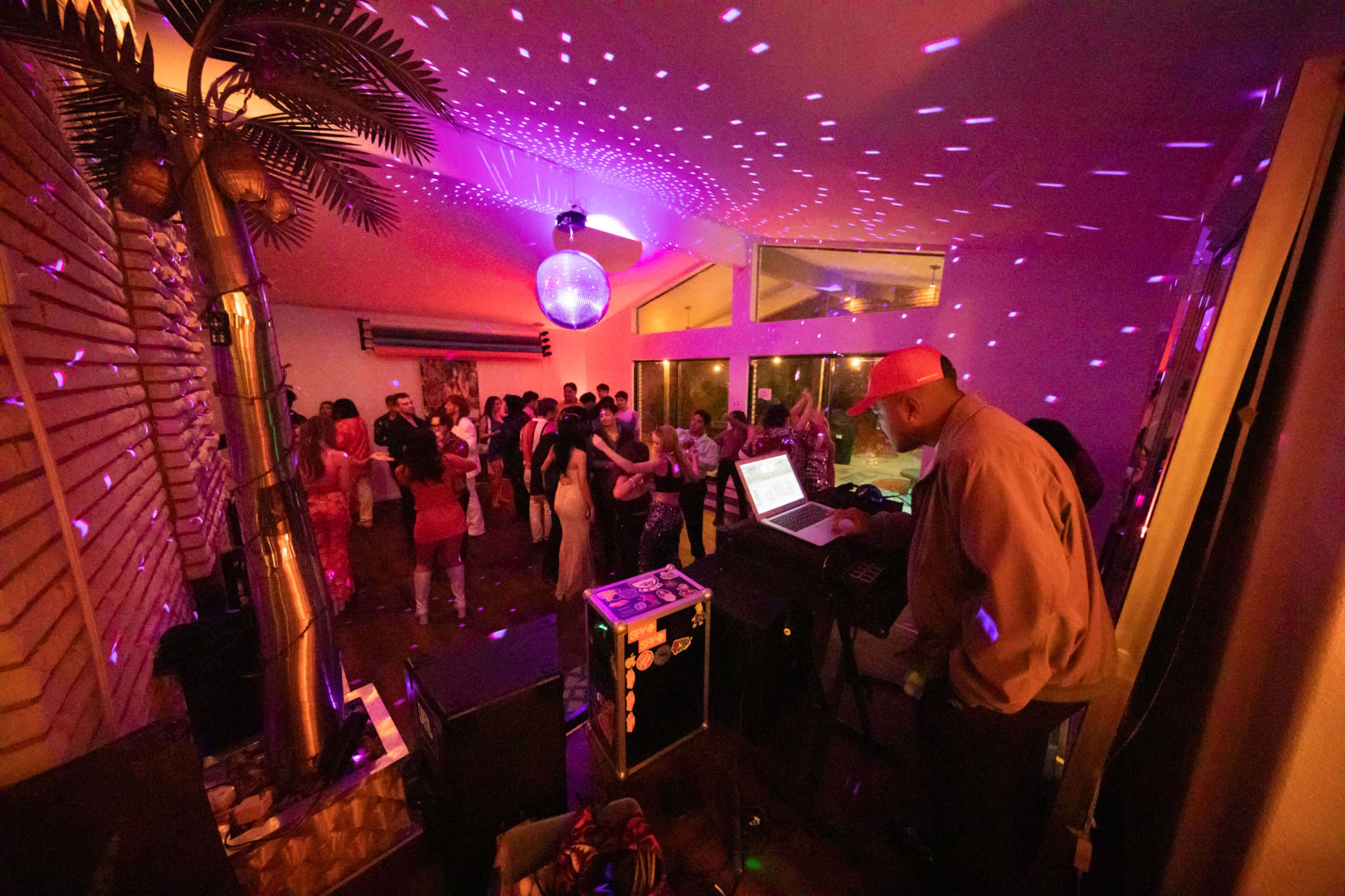 A DJ stands in front of a laptop while partygoers dance under colorful lights in a vibrant indoor setting.