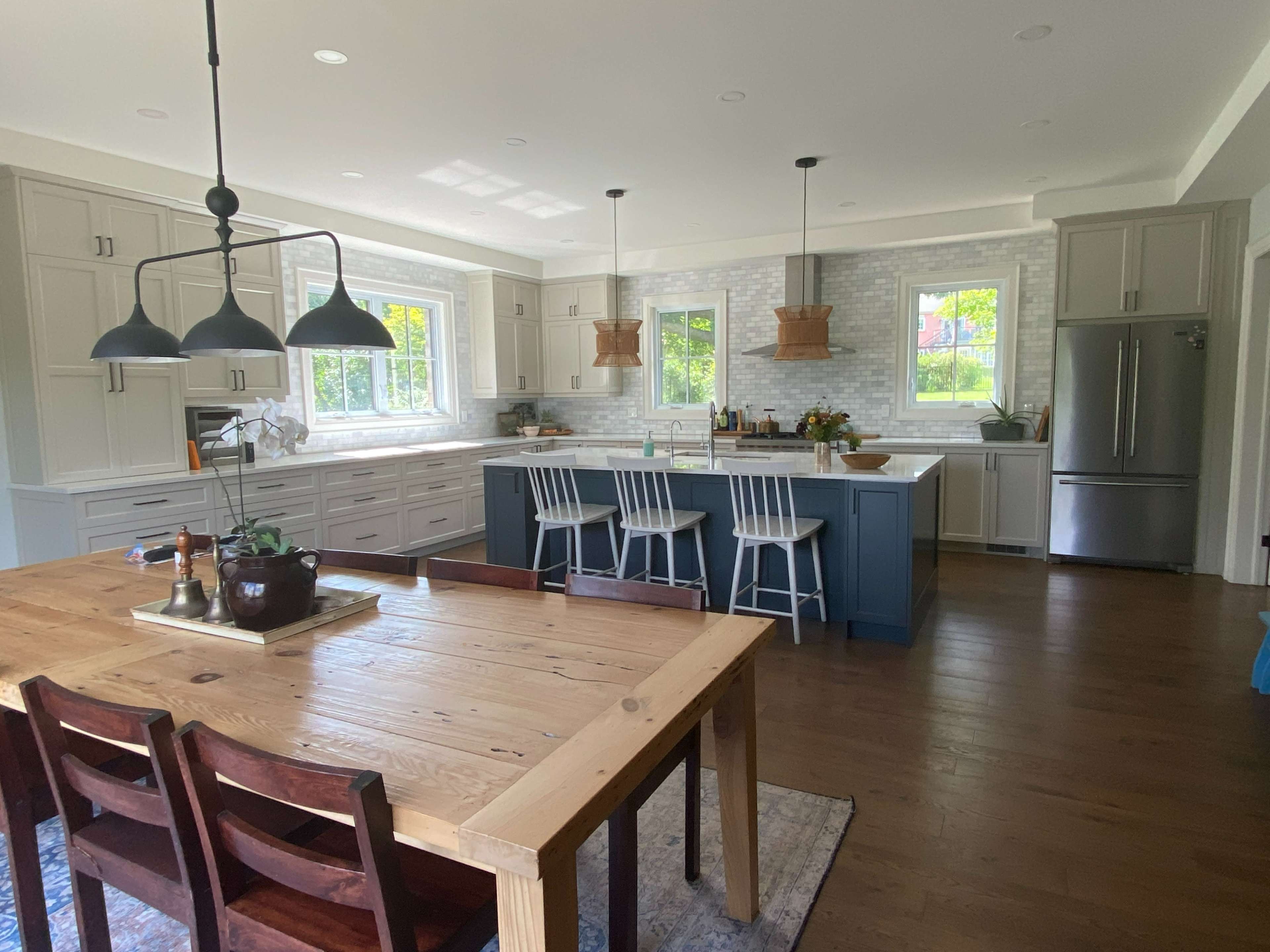A modern kitchen features light gray cabinetry, a central island with bar stools, and a wooden dining table, all set against a backdrop of large windows and natural light.