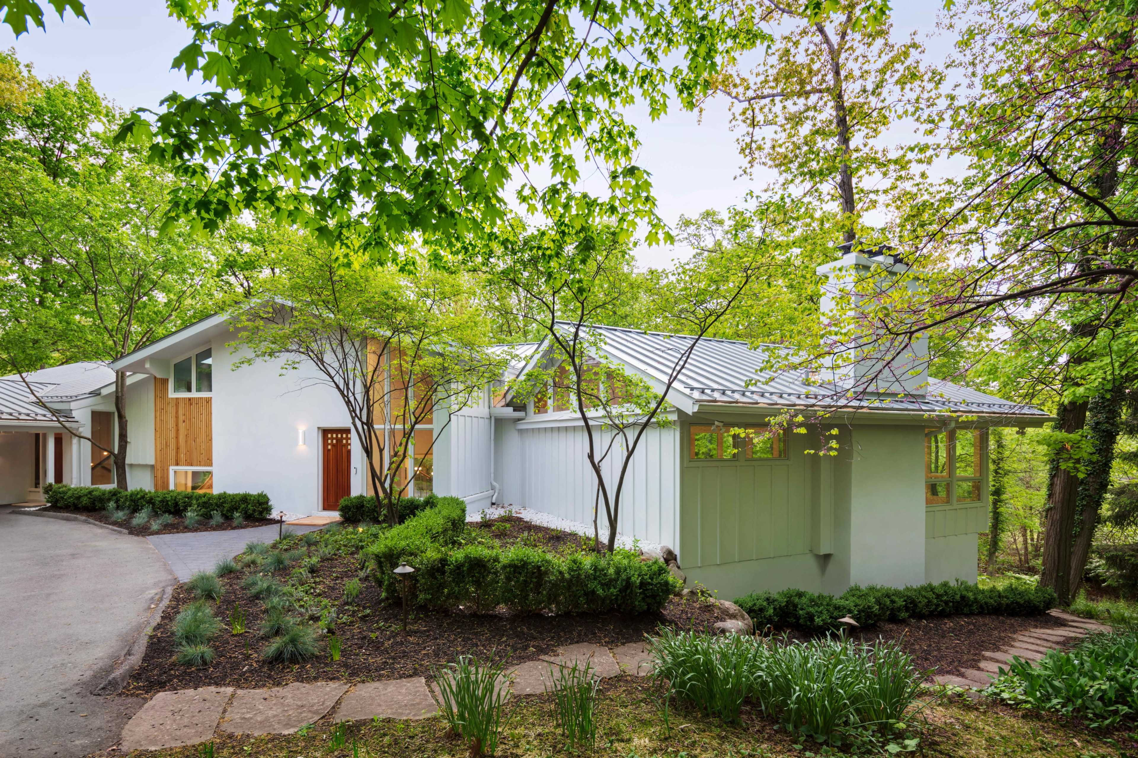 The image shows a modern, light-colored house surrounded by greenery and trees, with a driveway leading to the entrance.