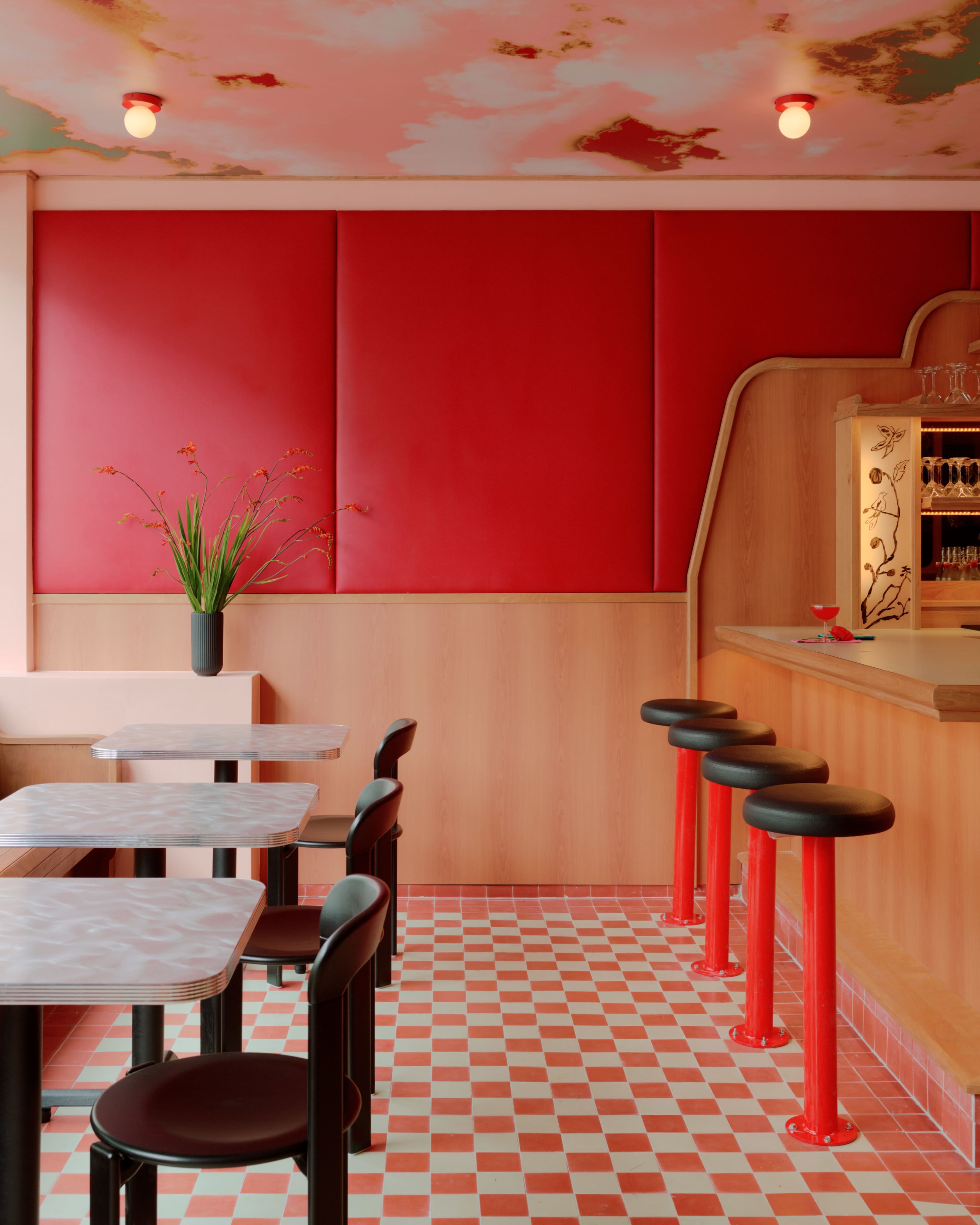 The interior of a modern bar features a red wall, wooden accents, and round black stools arranged around small tables on a checkered floor.