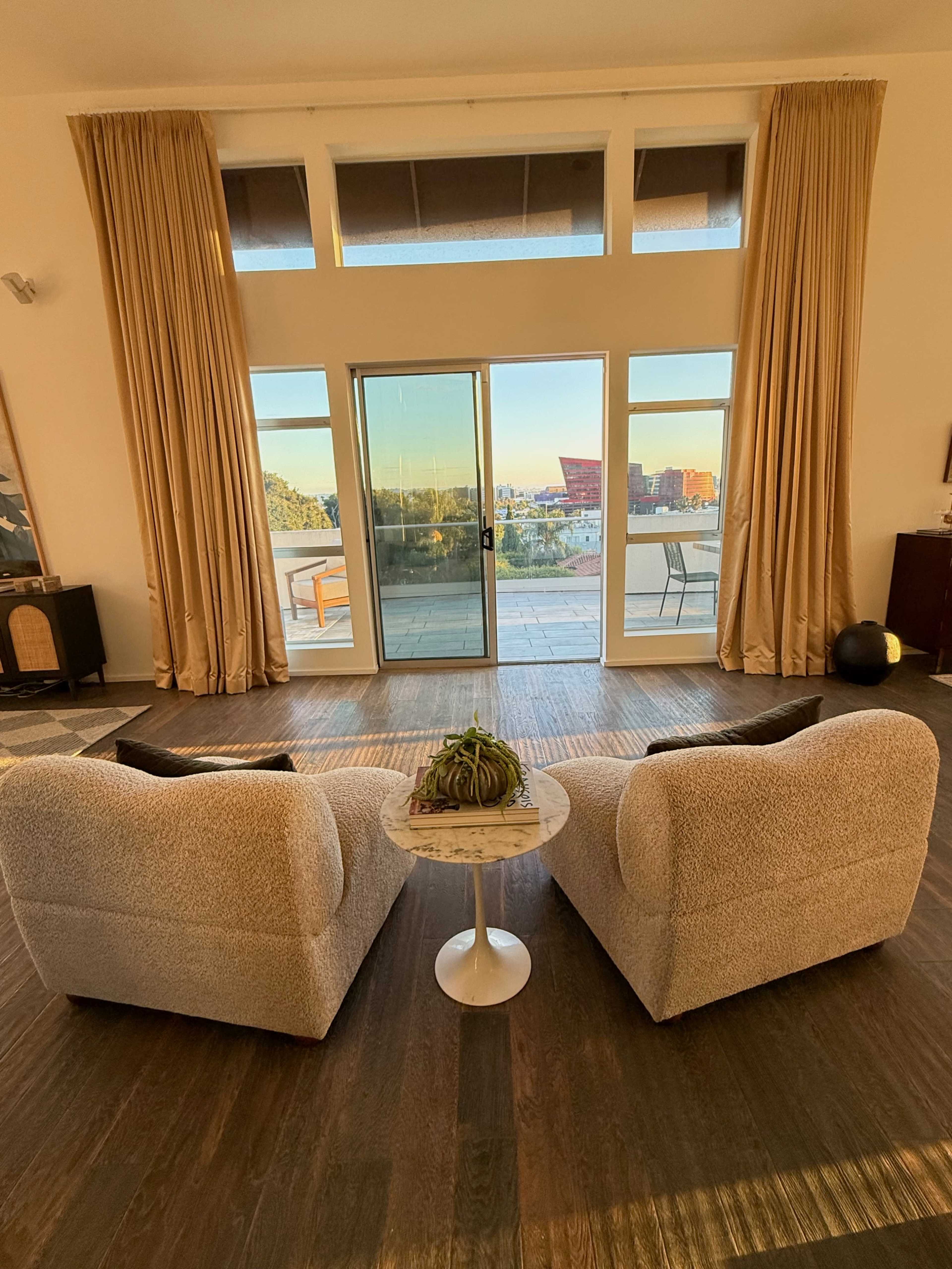 The image shows a modern living room with two upholstered chairs facing a small round table, overlooking large glass doors that open to an outdoor view.