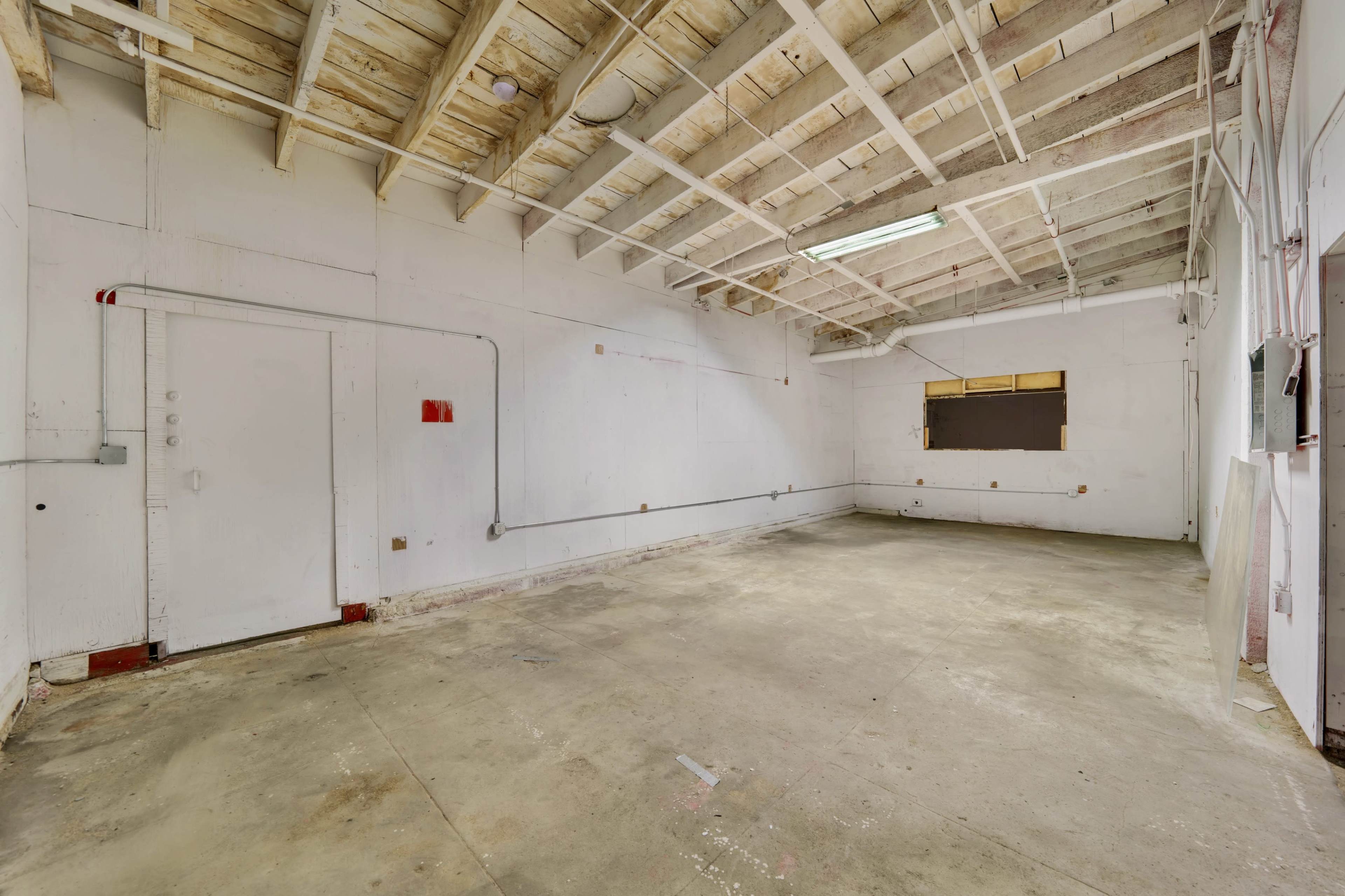 An empty room with bare concrete flooring, white walls, exposed beams, and a small window.
