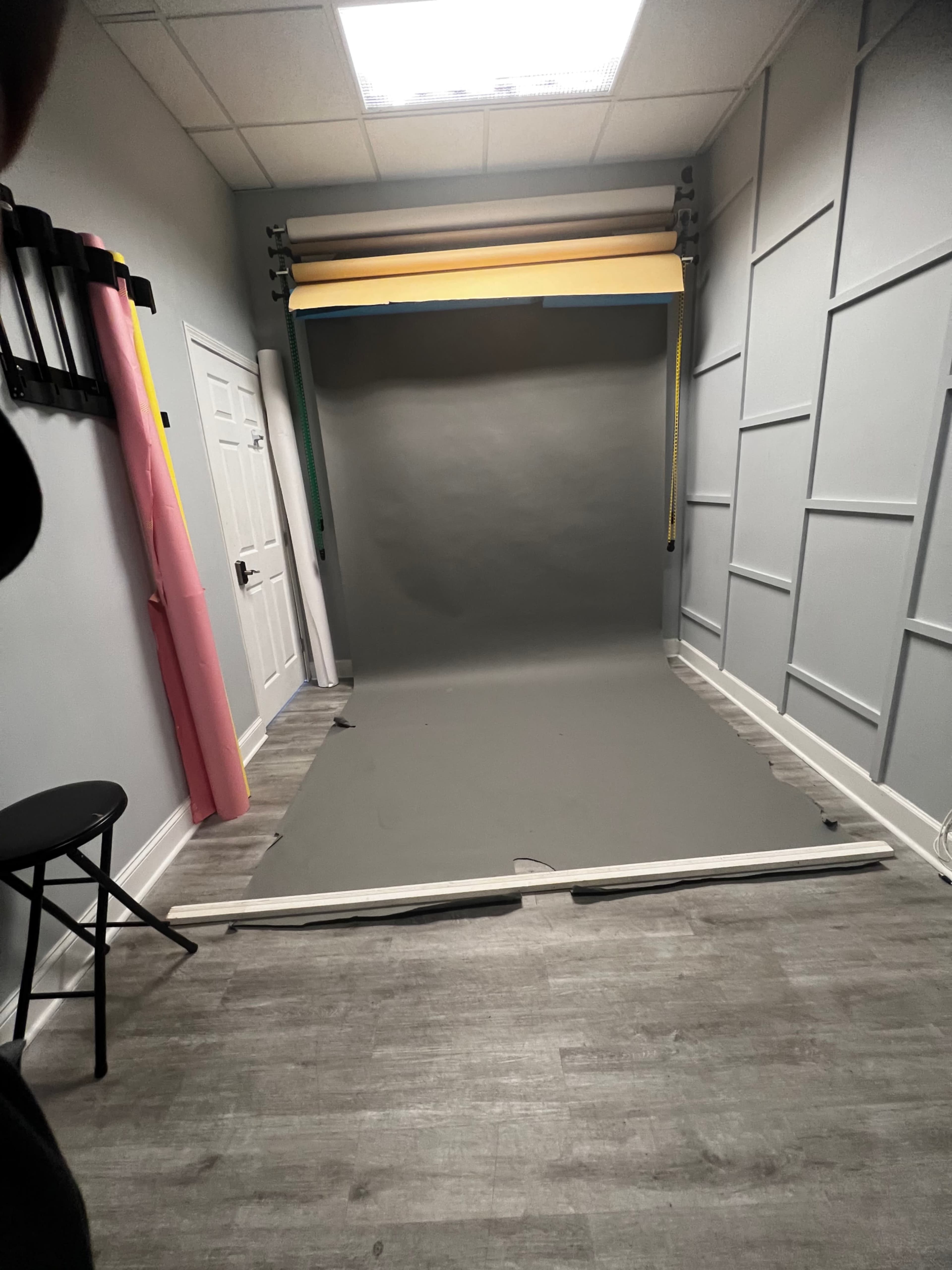 A photography studio with a gray backdrop set up against a wall, flanked by colorful paper rolls and a chair in the corner.