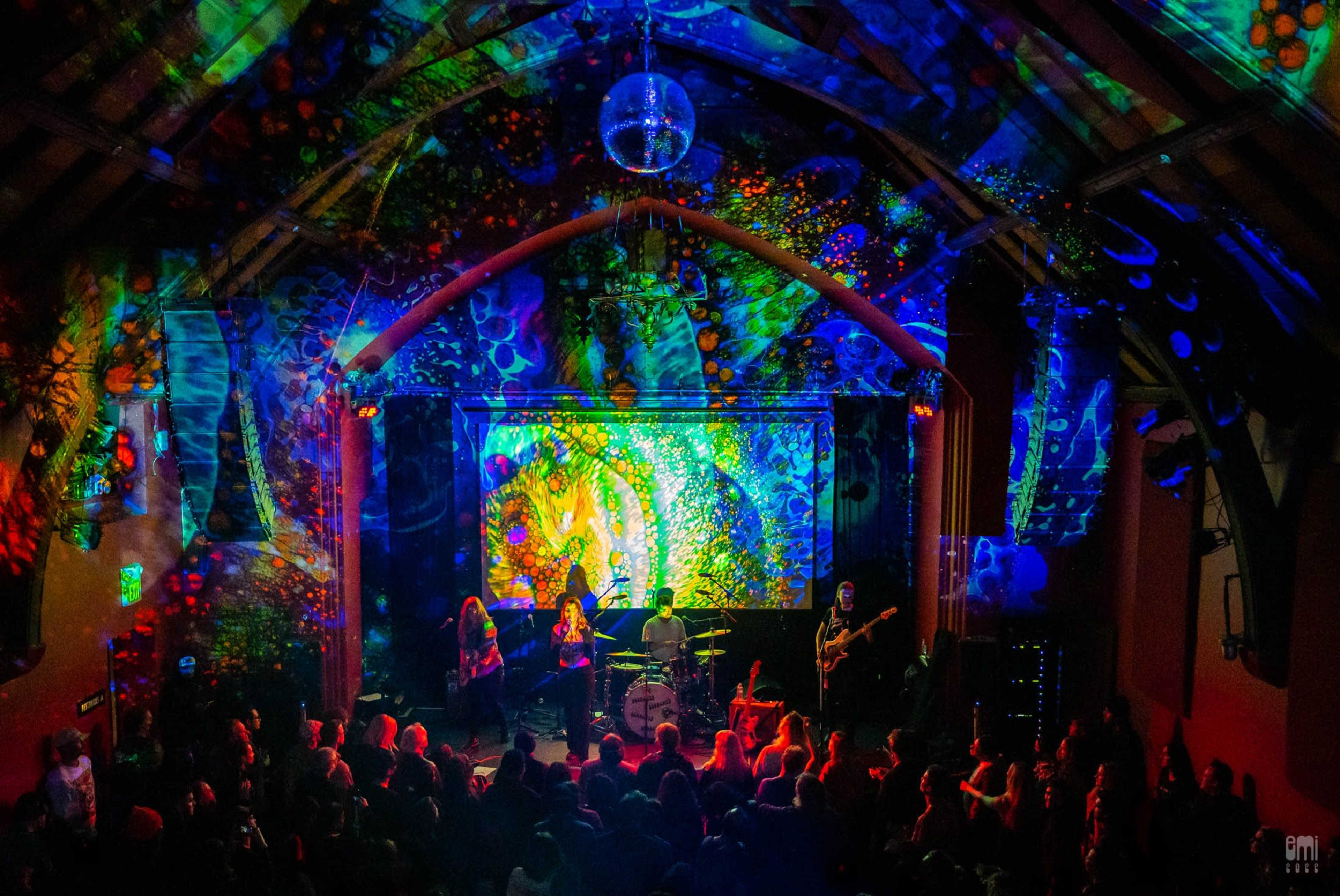 A band performs on stage in a vibrant, illuminated venue with colorful projections on the walls and ceiling.