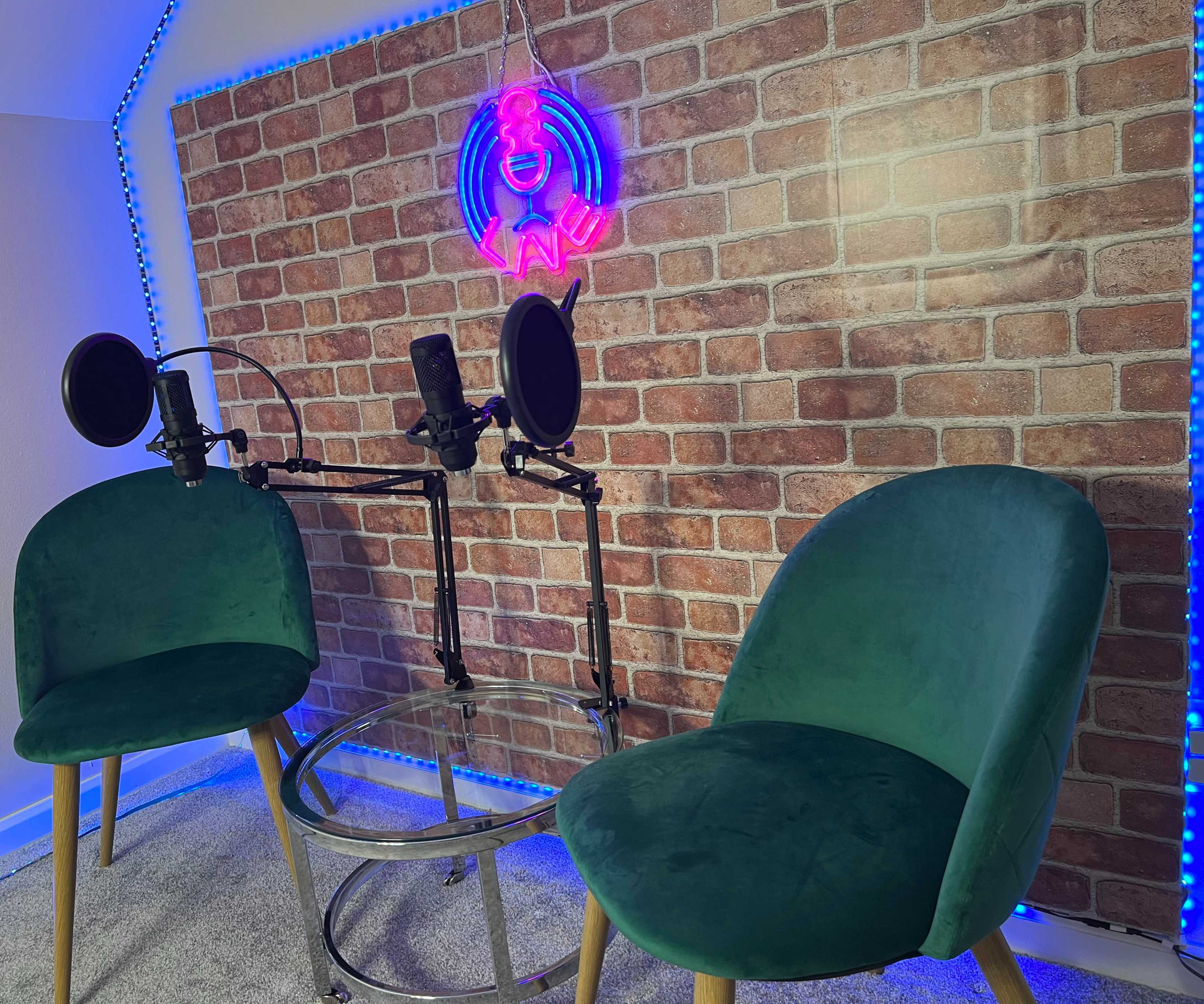 The image shows a podcasting setup featuring two green chairs, a round glass table, and two microphones positioned on adjustable stands against a brick-patterned background with neon lighting.