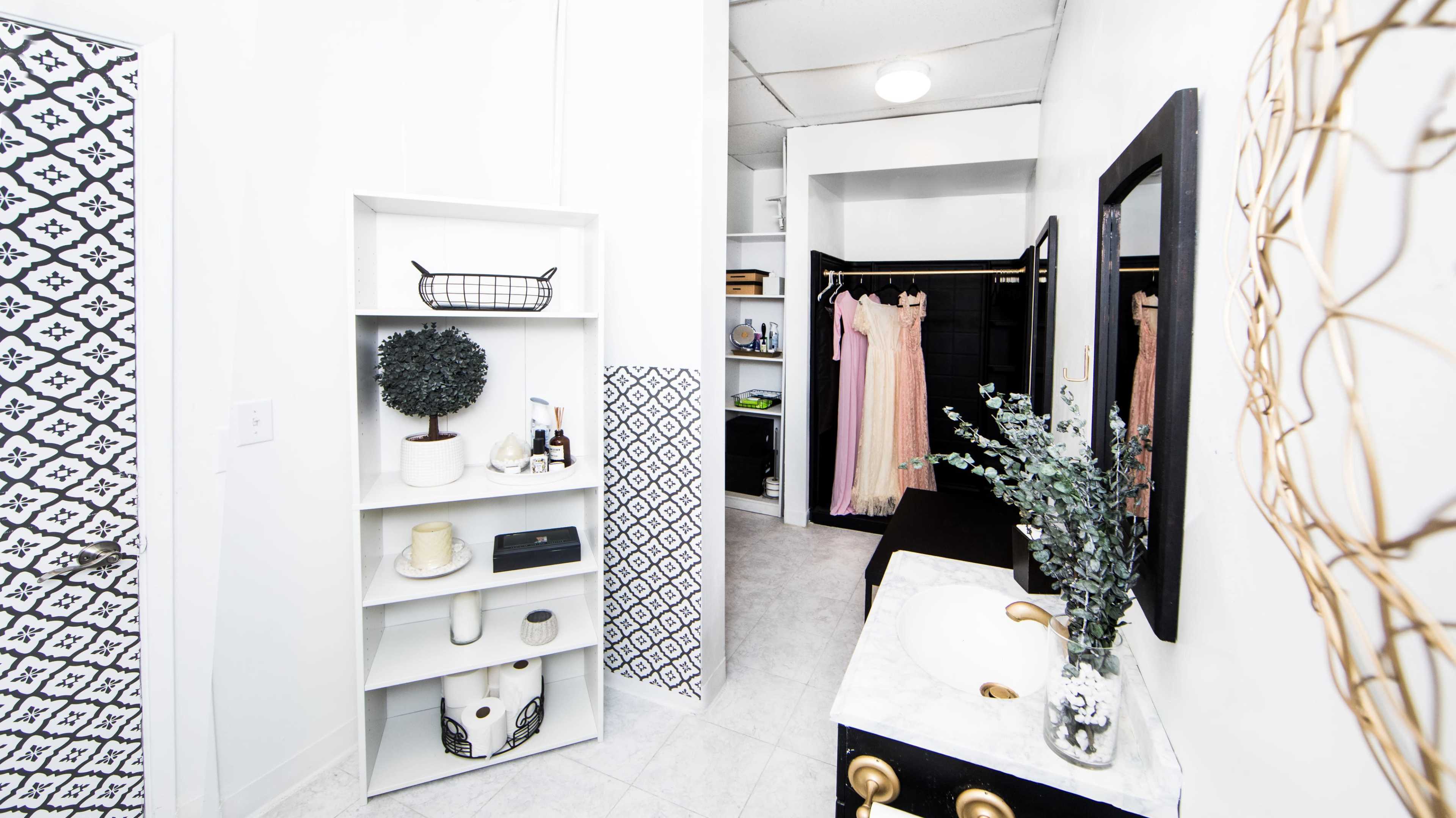 The image shows a modern bathroom with a white color scheme, featuring a small shelving unit, a vanity with a sink, and a closet with hanging garments.