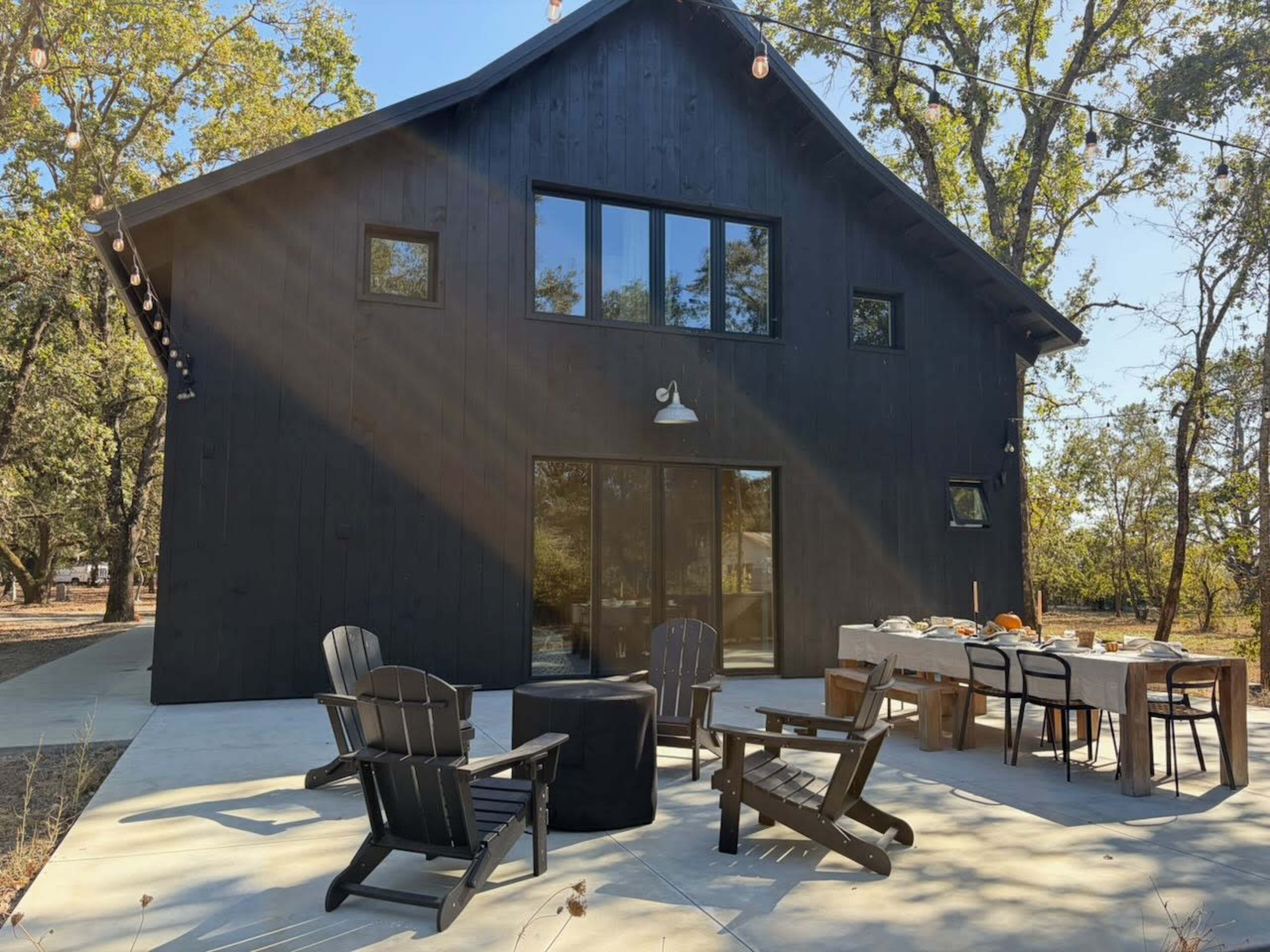 A modern black farmhouse with large windows and a patio set up for dining sits amidst trees in a sunny outdoor setting.